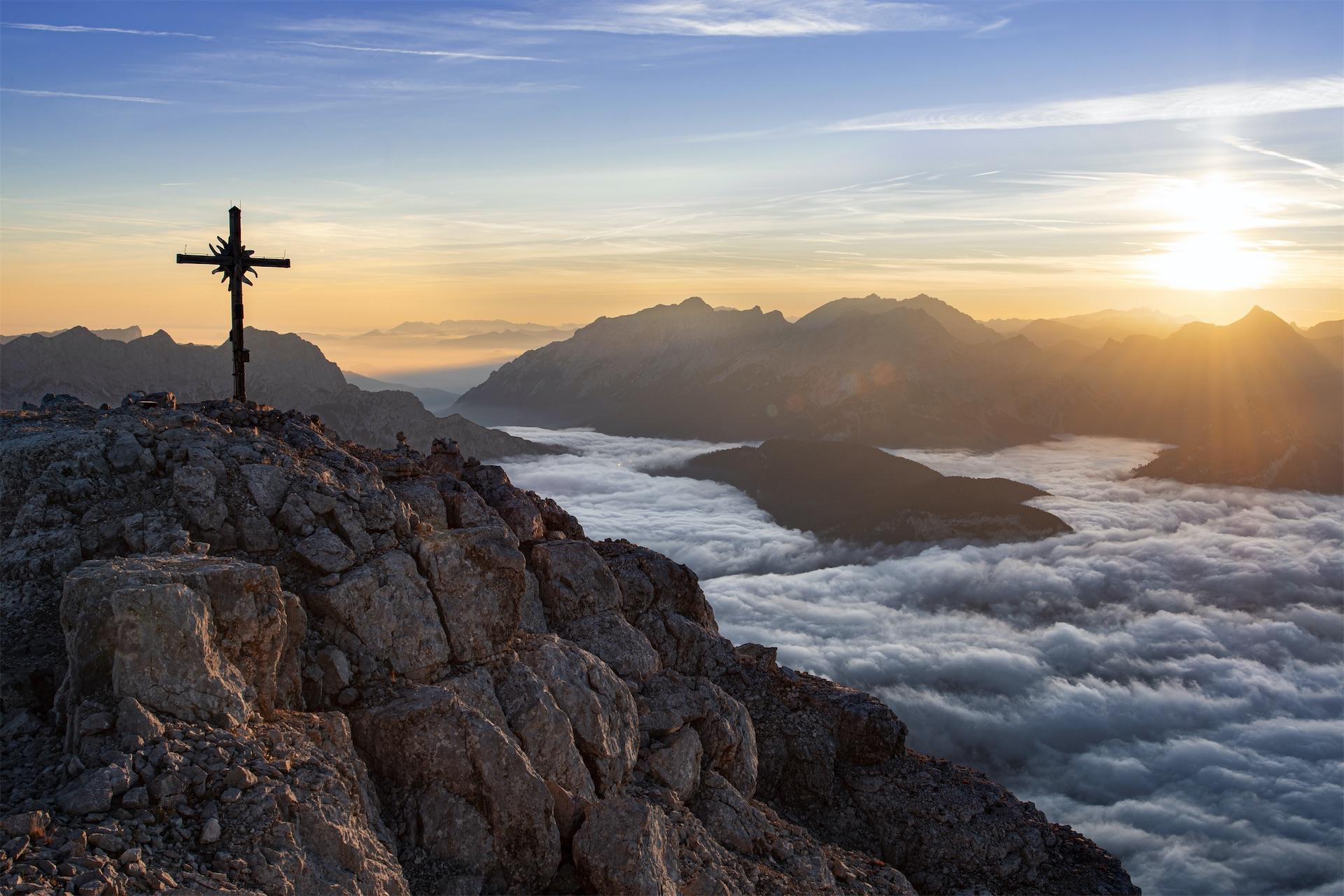 A summit cross atop a rocky mountain peak overlooking a sea of clouds at sunrise.