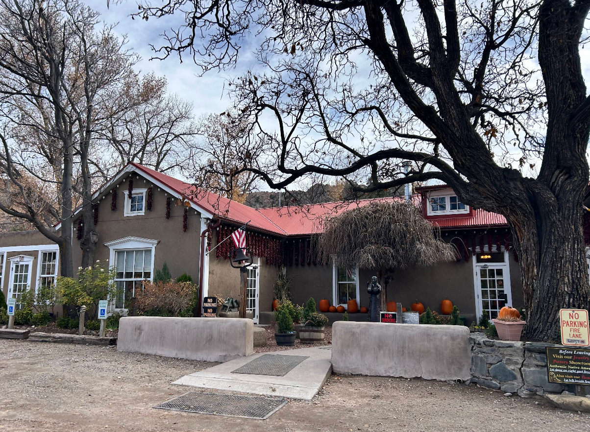An adobe-style building with a red metal roof, decorated with strings of red chili peppers, featuring pumpkins on the porch and two handicap parking signs in the foreground.