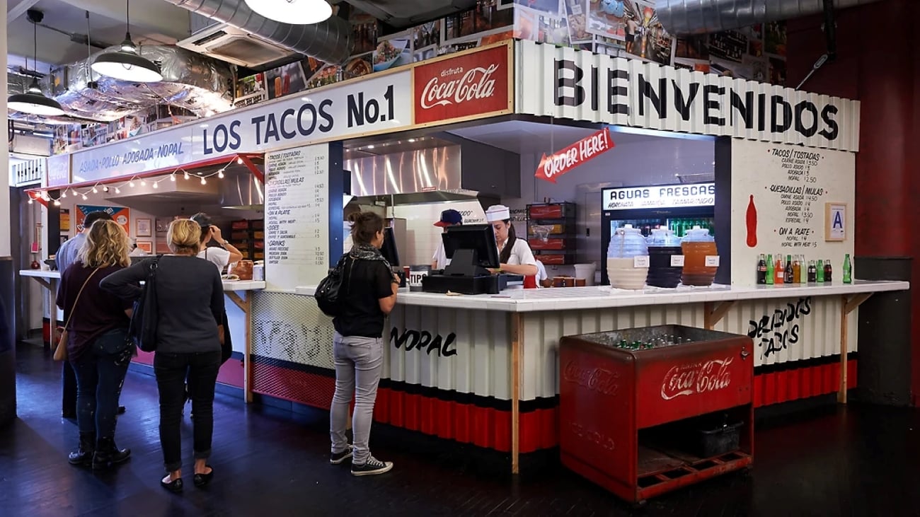 Customers stand at the counter of a Los Tacos No. 1 restaurant, where a printed menu is displayed above the ordering area and a red vintage Coca-Cola crate sits in the foreground.