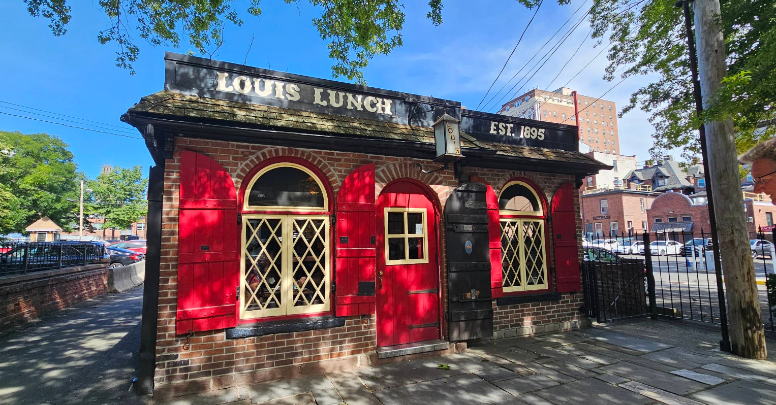A small, historic red brick building with yellow-trimmed windows and a black “Louis Lunch Est. 1895” sign—one of the Restaurants That Still Cook over classic methods—surrounded by trees and city buildings under a blue sky.