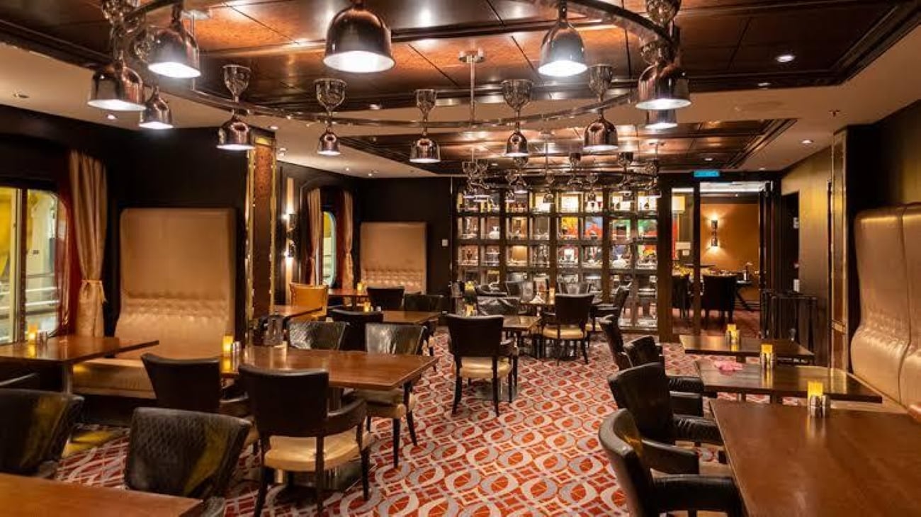 The interior of a dimly lit, upscale restaurant featuring a circular chandelier over dining tables with dark chairs and patterned carpeting.