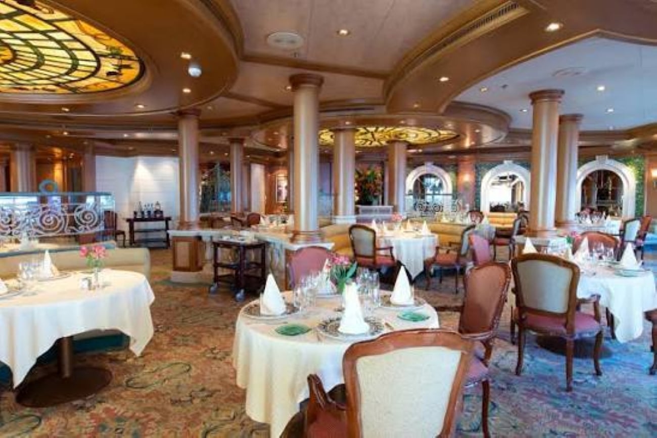 Cruise ship specialty restaurants ad fine dining featuring round tables with white tablecloths and patterned carpet.