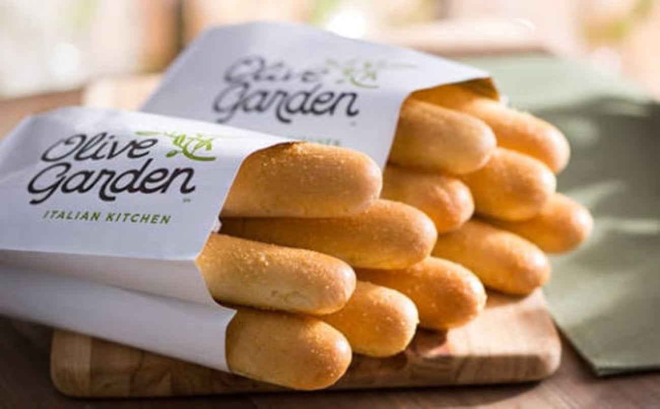 Two paper bags filled with Olive Garden breadsticks sitting on a wooden cutting board.