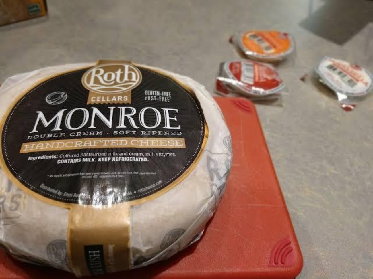 A round wheel of Roth Cellars Monroe soft ripened cheese on a red cutting board, considered the best souvenir, with three small, circular plastic containers in the background.