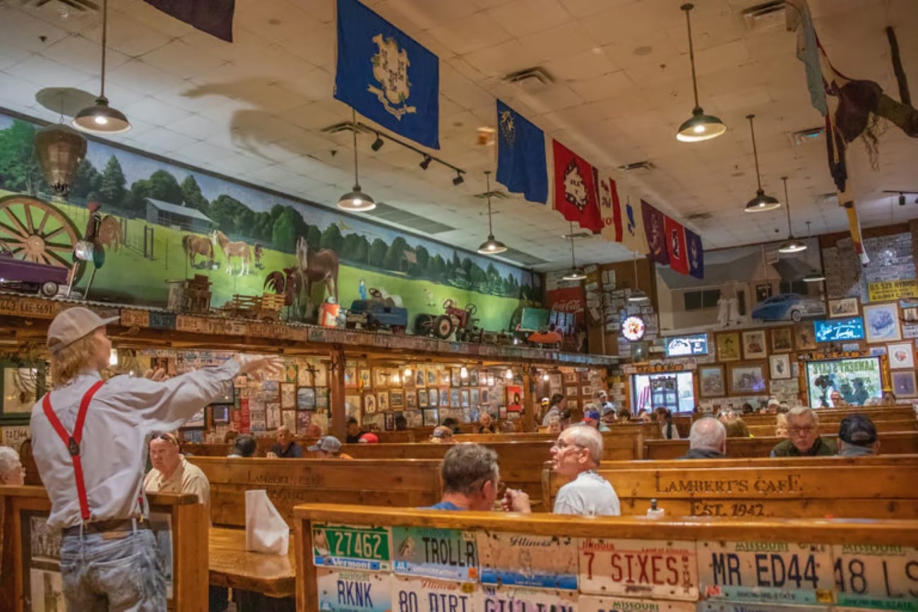 A server in suspenders tosses a bread roll to a customer inside a restaurant decorated with license plates and a mural of a countryside scene.