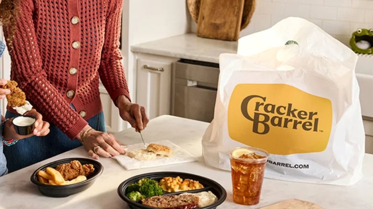 A person holding a piece of fried chicken over a container, next to takeout containers of meat and sides, a glass of iced tea, and a Cracker Barrel bag.
