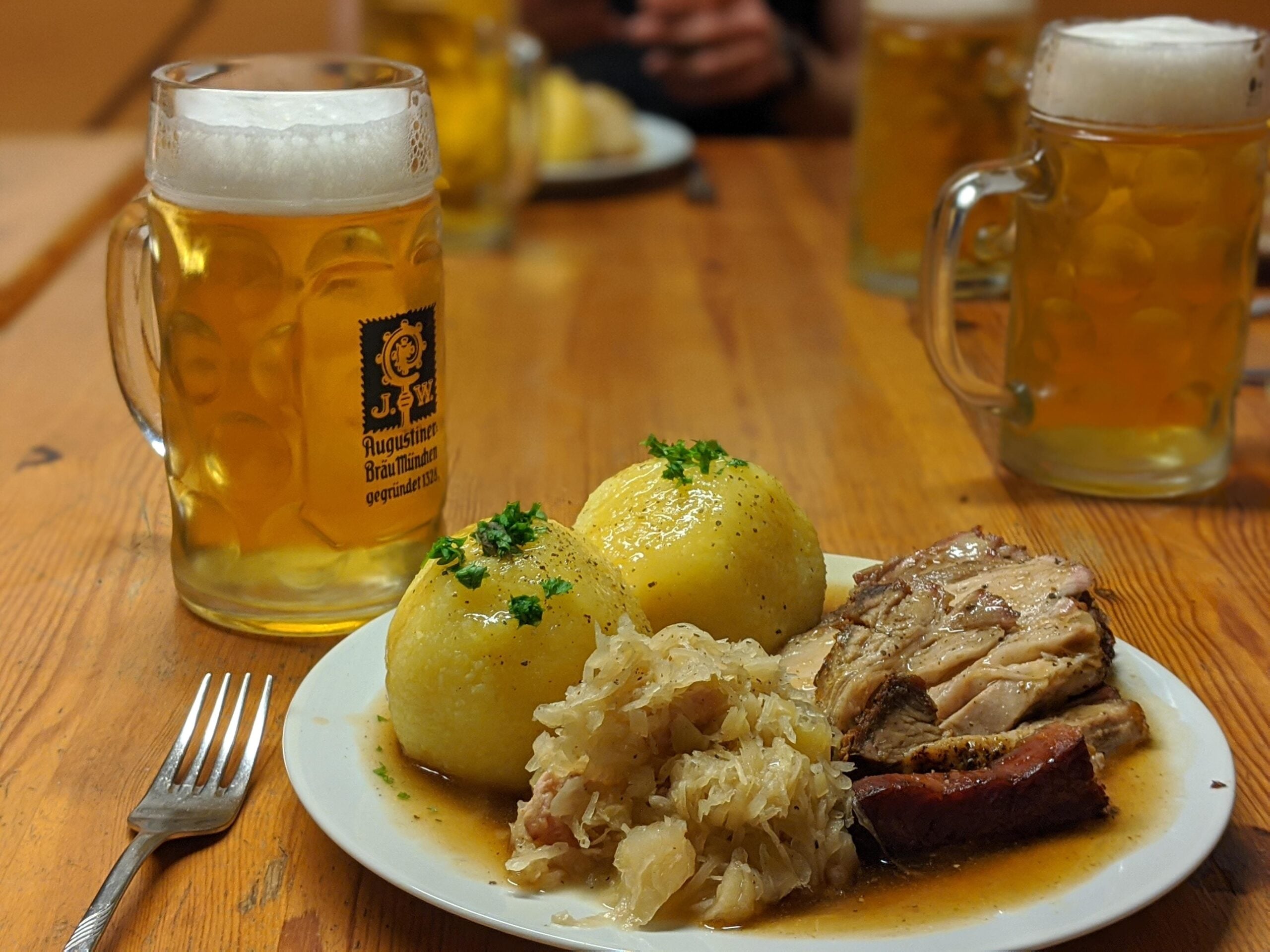 A plate with two potato dumplings, sauerkraut, and roasted pork, served alongside a glass beer mug on a wooden table.