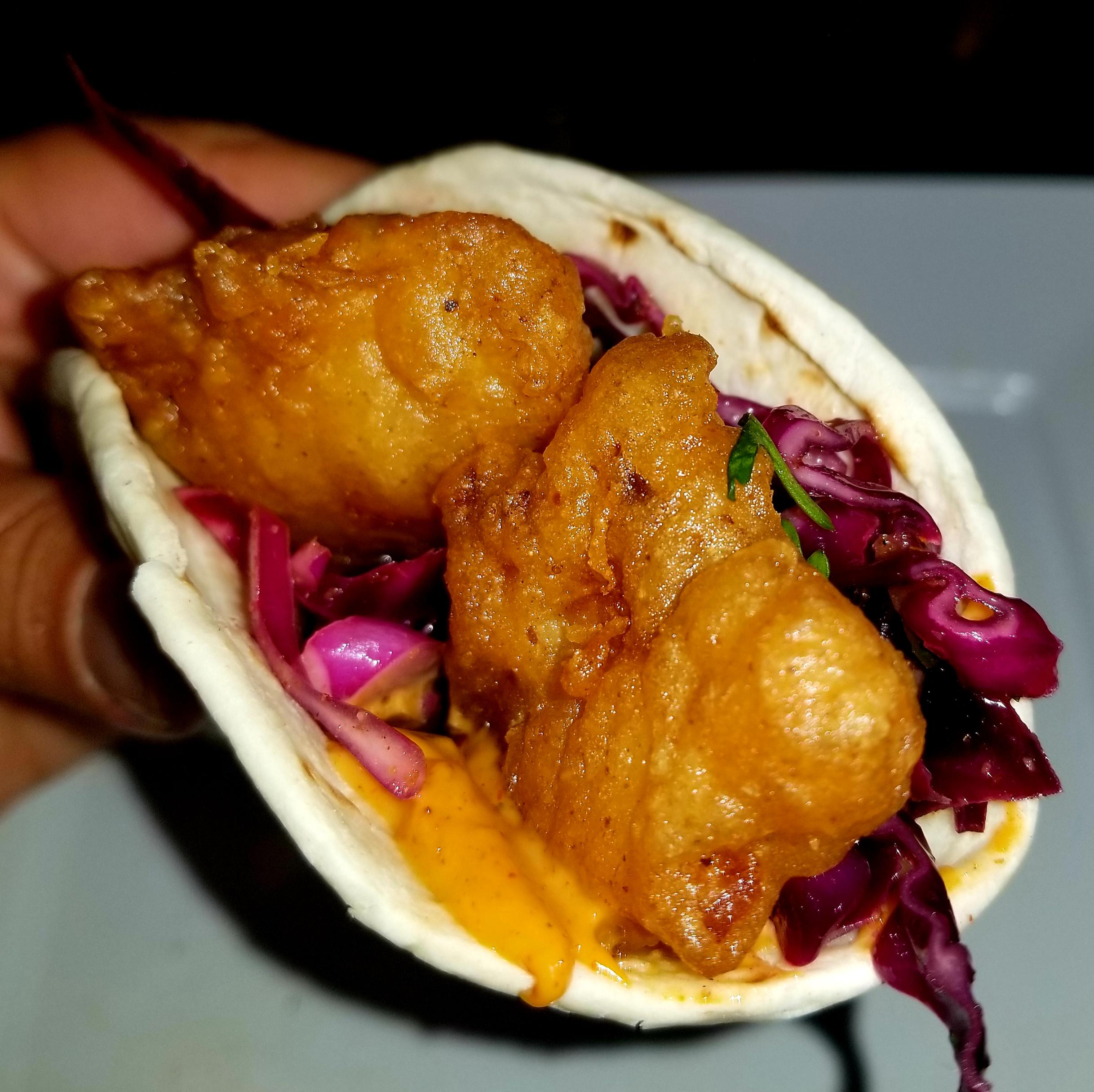 A hand holds a fried fish taco in a flour tortilla with red cabbage slaw and orange sauce.