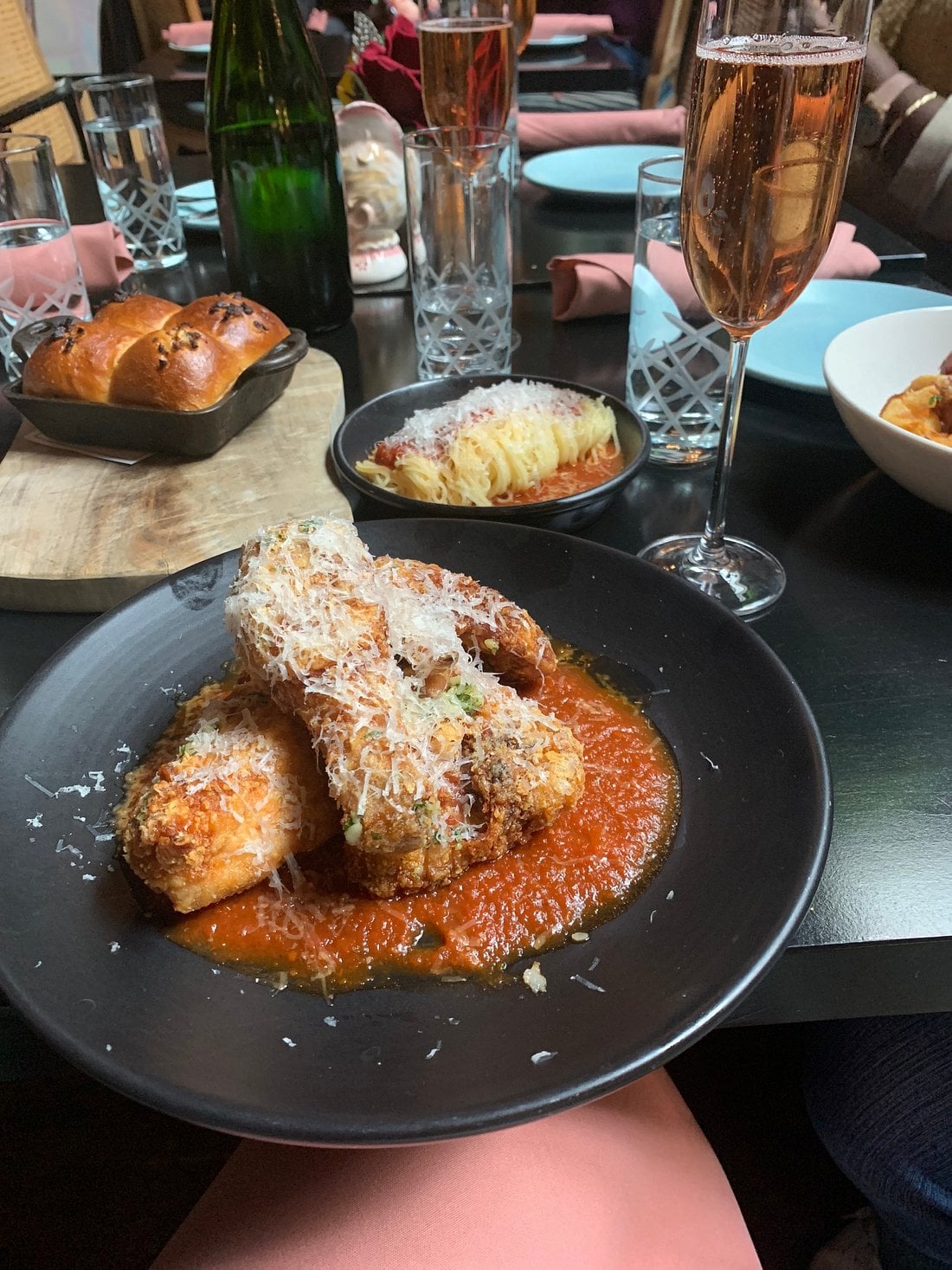 A black plate featuring breaded chicken cutlets topped with grated cheese in a pool of red sauce, with a small bowl of pasta and bread rolls on the table nearby.