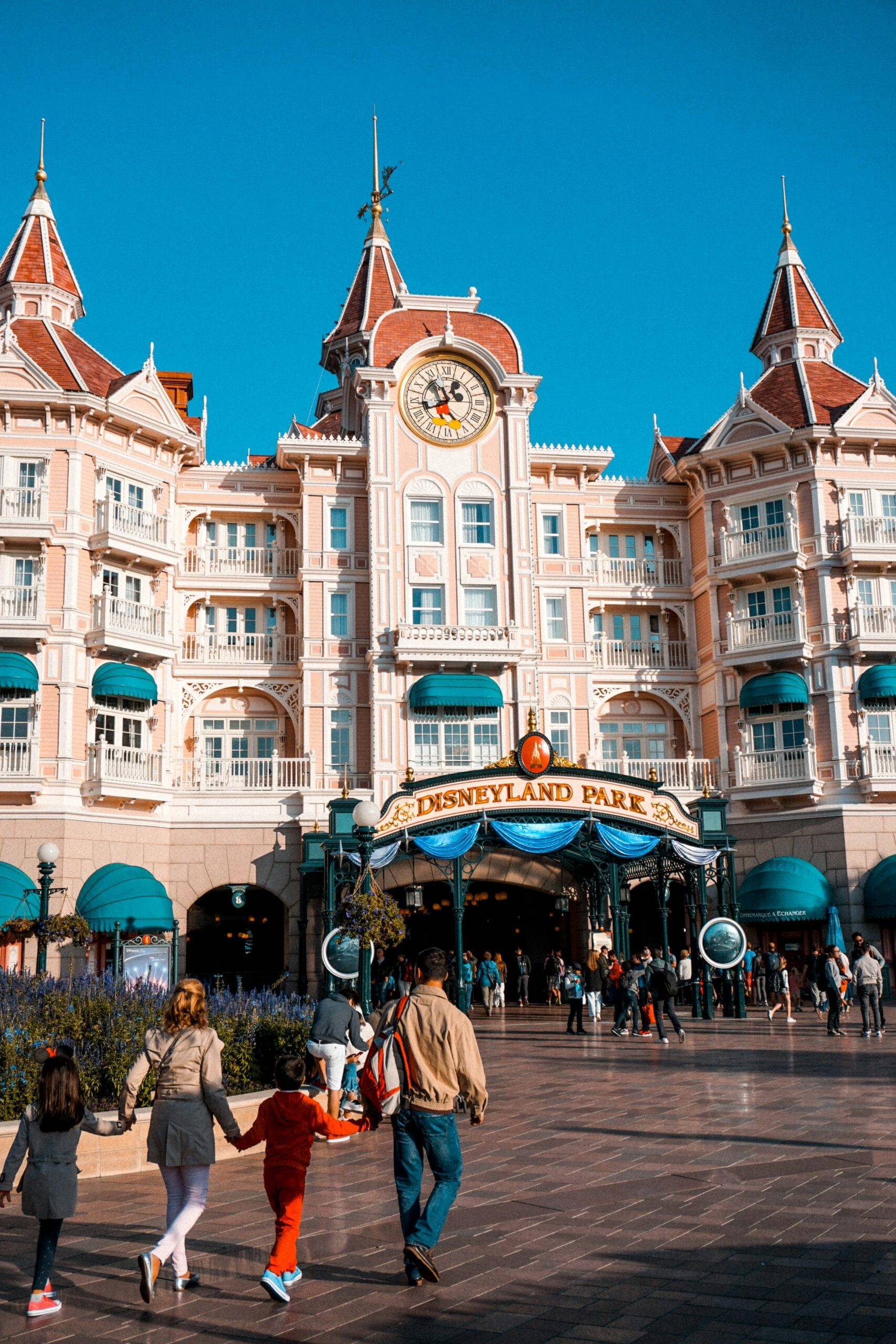 Families walk toward the entrance of Disneyland Park, in one of these family trips, located beneath a grand pink Victorian-style building with a clock and turrets under a clear blue sky.