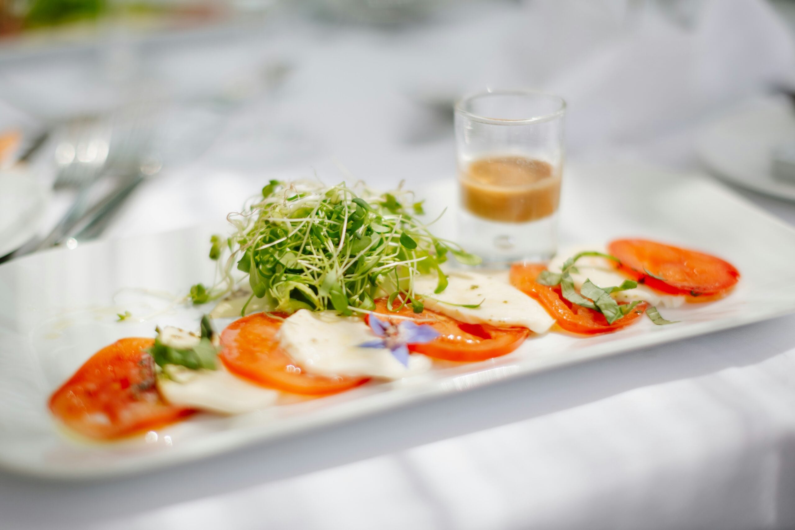 Caprese salad with sliced tomatoes, mozzarella, microgreens, and a small glass of dressing on a rectangular plate.