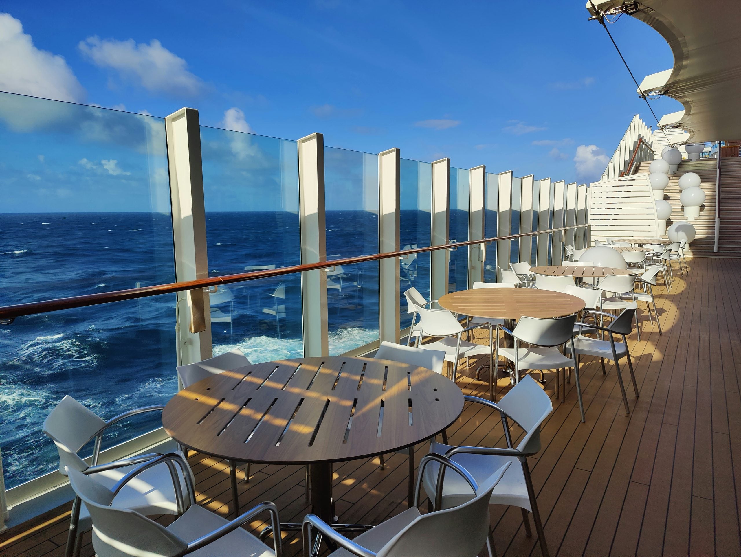 Sunny outdoor deck on a cruise ship with round wooden tables, white chairs, glass railing, and an ocean view. Stairs lead up, and decorative lights hang above. Waves and blue sky are visible in the background.