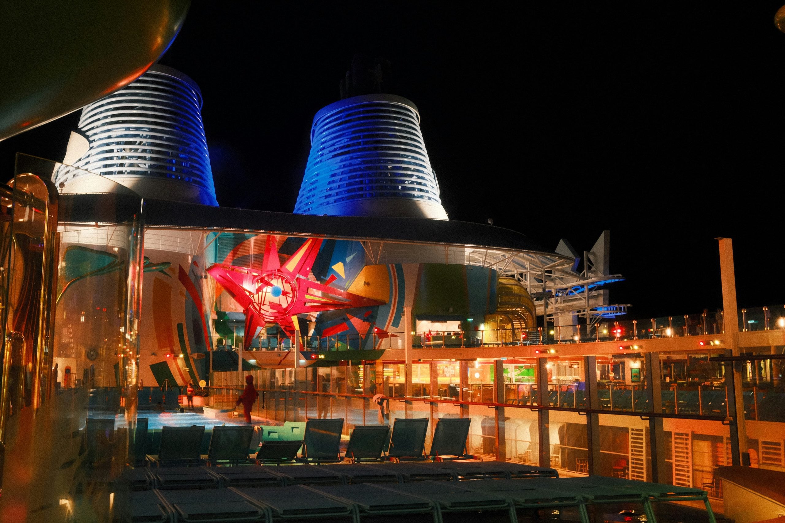 A brightly lit cruise ship deck at night features colorful abstract wall art, empty lounge chairs, and illuminated smoke stacks, with a few people walking in the background.