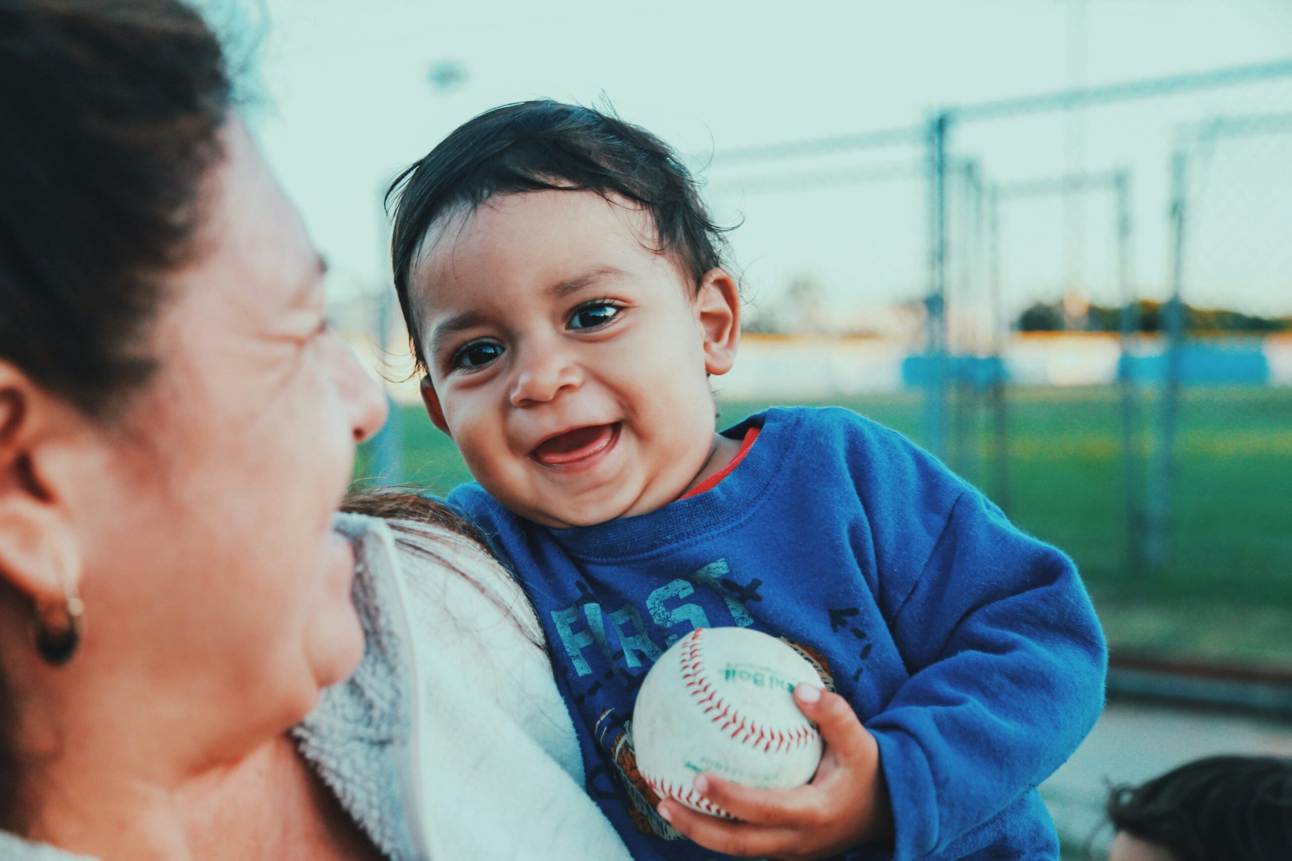A smiling baby in a blue sweatshirt holds a baseball while being held by an adult woman at an outdoor sports field.