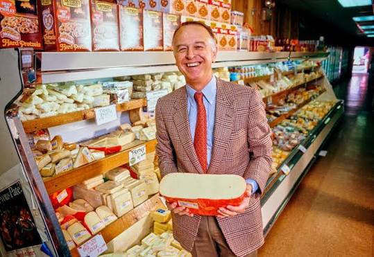 A smiling man in a blazer holds a large, round block of cheese in front of a store deli counter filled with various cheeses and snacks. Trader Joe’s facts.