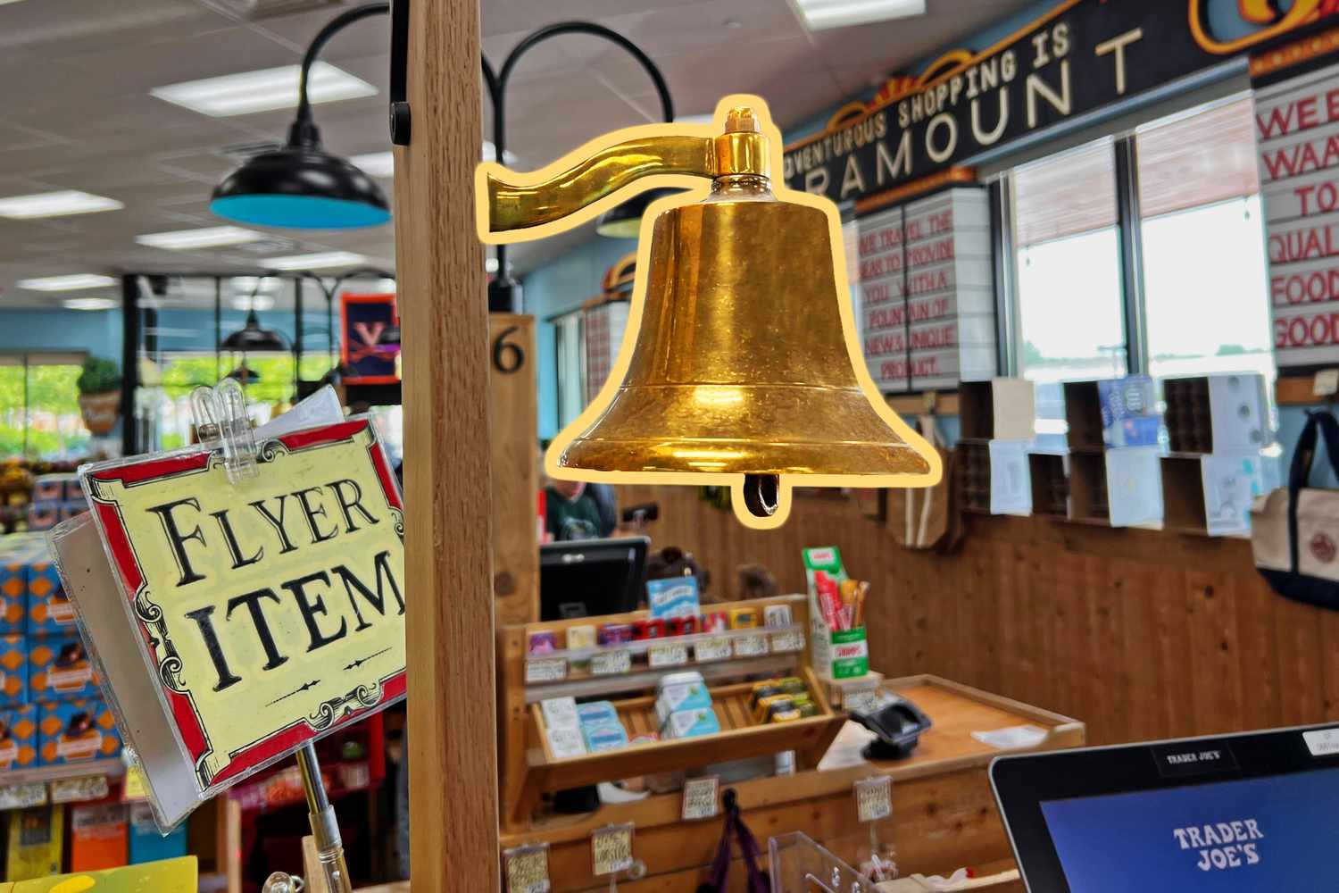 A brass bell mounted on a wooden display inside a grocery store, next to a sign that reads “Flyer Item.”