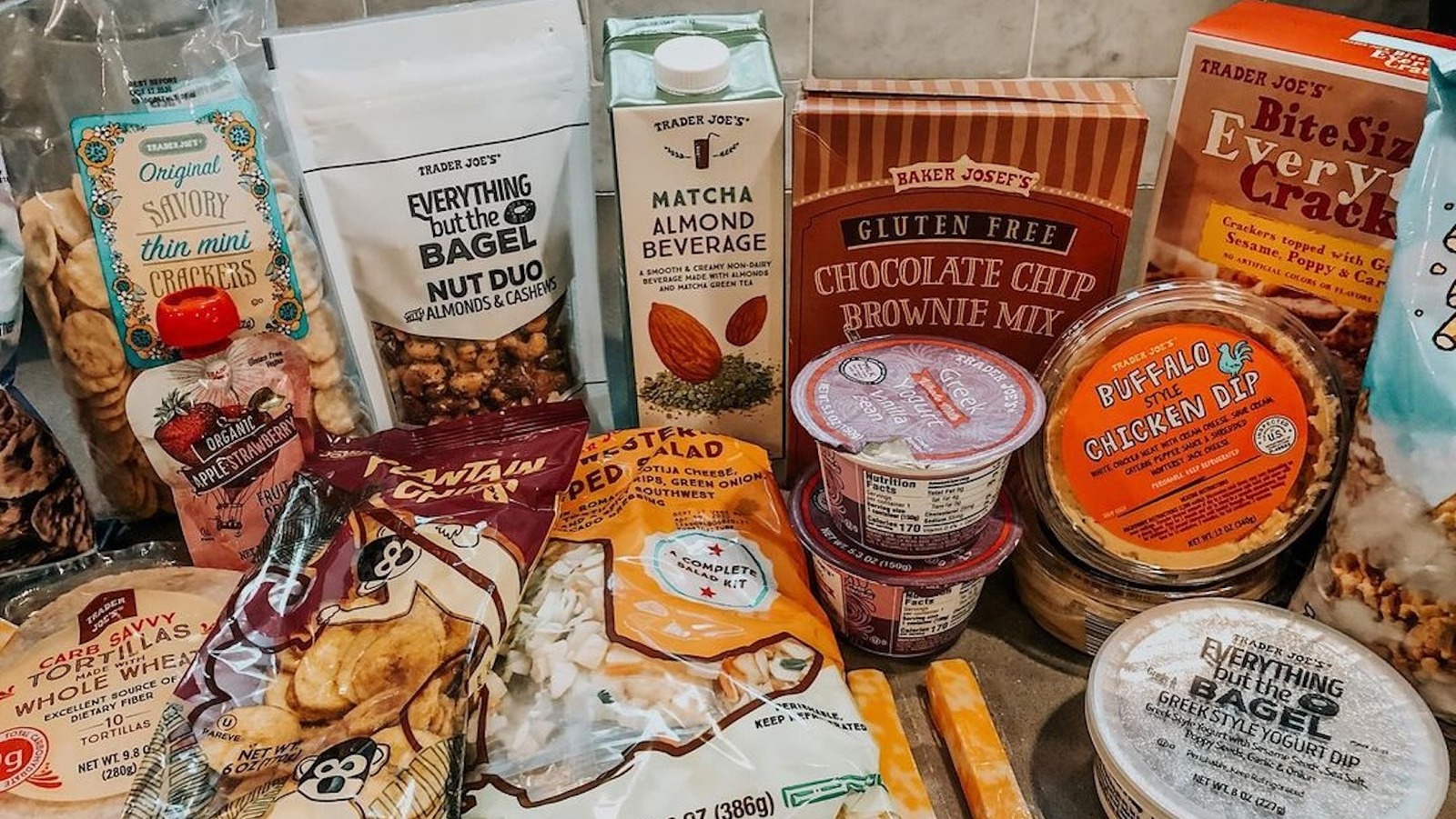 An assortment of Trader Joe’s brand food products including crackers, nuts, matcha almond beverage, brownie mix, greek yogurt, buffalo chicken dip, plantain chips, a salad kit, tortillas, and yogurt dip.