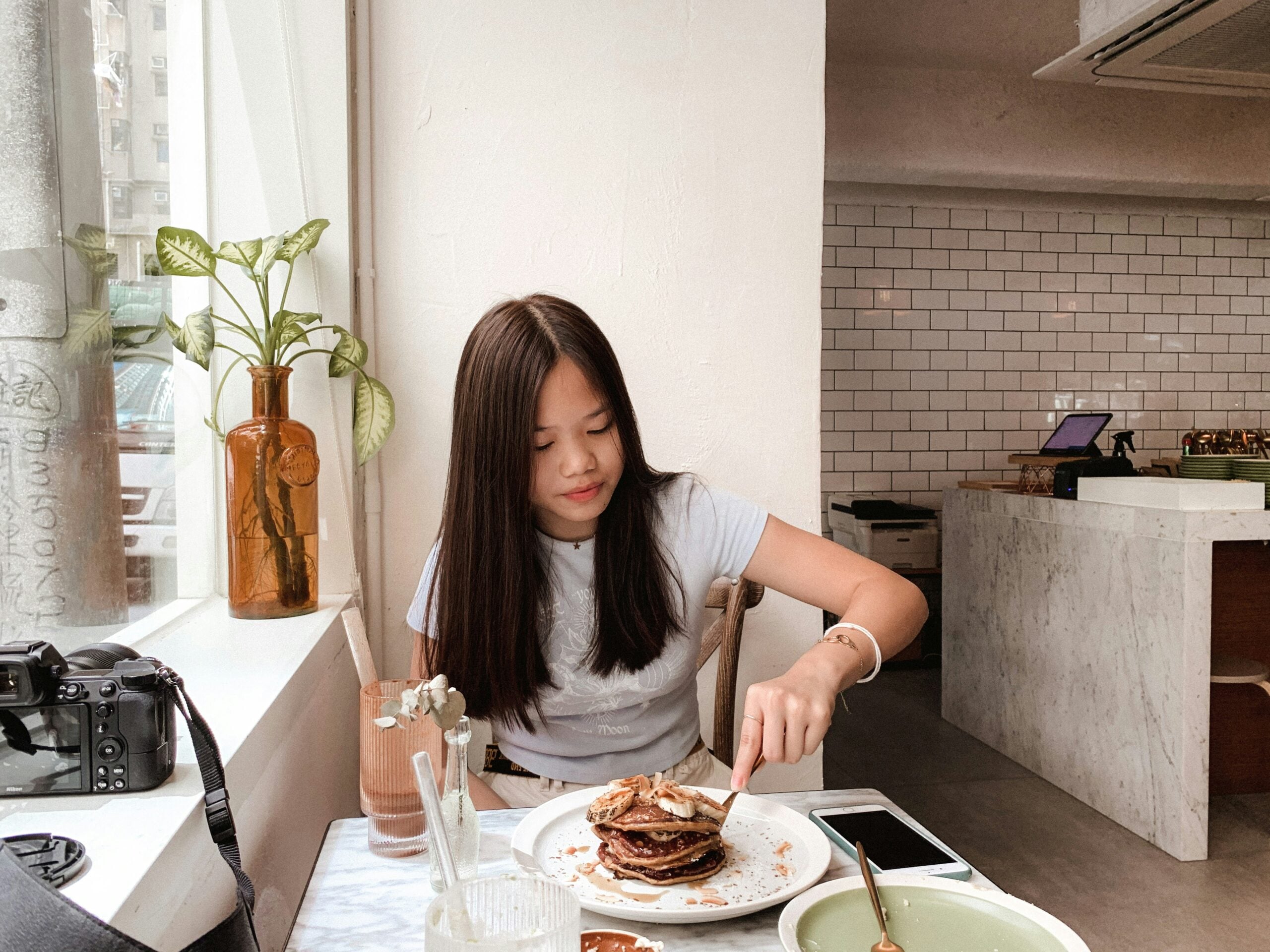 A young woman sitting at a table in a cafe cuts into a stack of pancakes topped with banana slices.