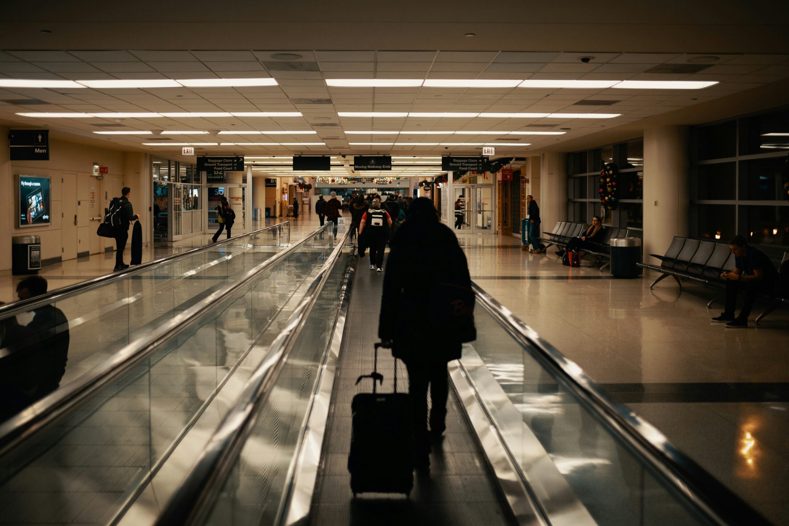 A person pulls a rolling suitcase while walking along a moving walkway in an airport terminal.