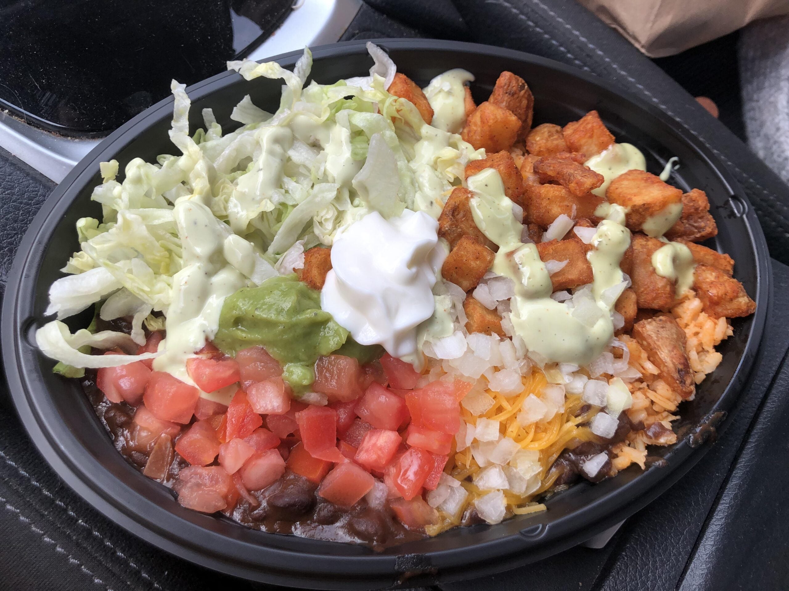 A black plastic bowl containing a burrito bowl with seasoned potatoes, black beans, rice, shredded lettuce, diced tomatoes, guacamole, sour cream, shredded cheese, and a creamy green sauce.