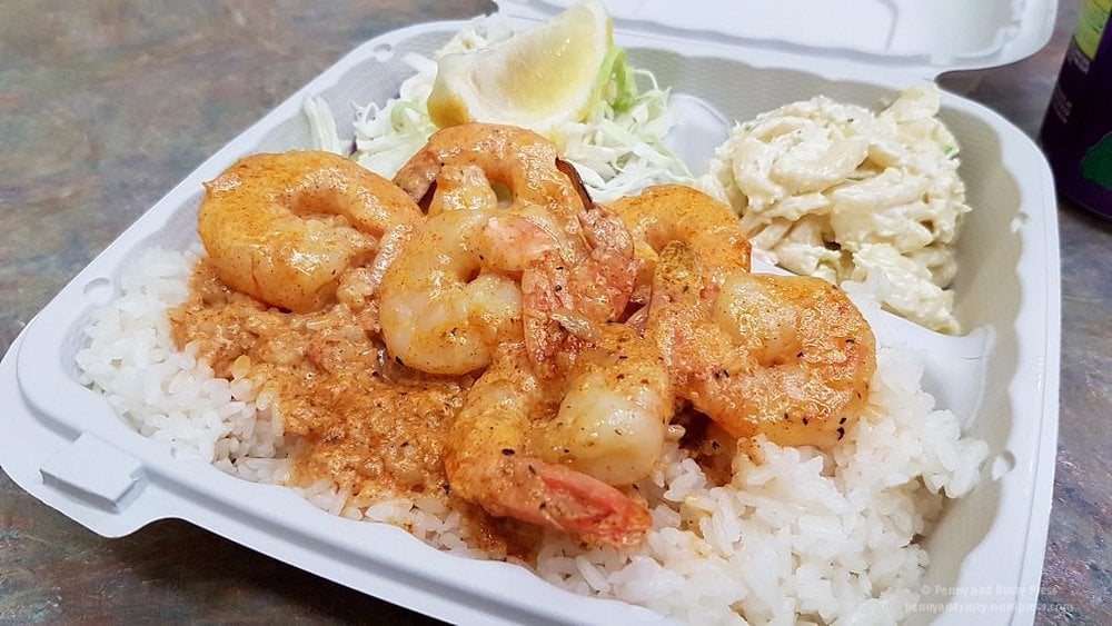 A takeout container with shrimp in a creamy sauce over white rice, served with macaroni salad, shredded cabbage, and a lemon wedge.