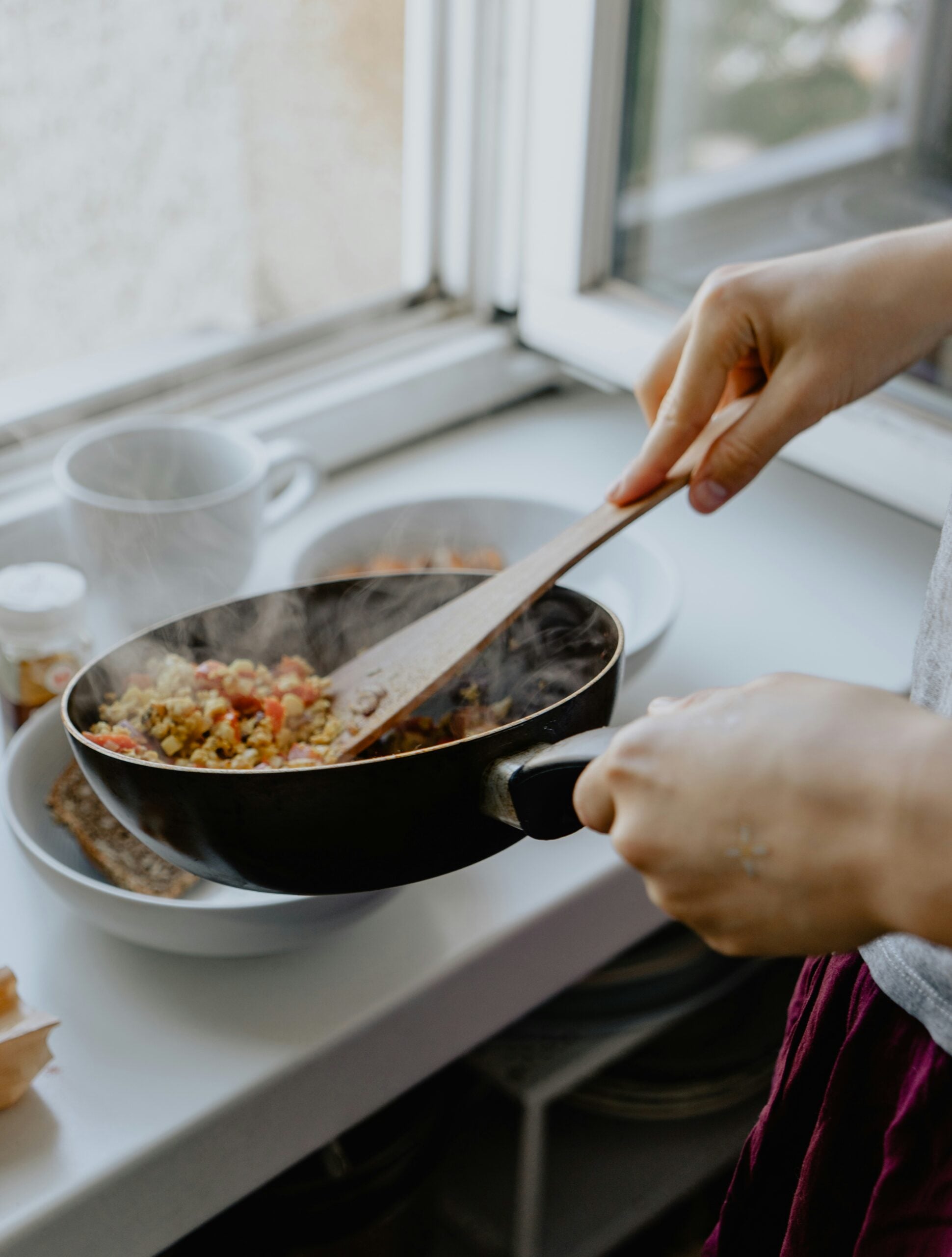 A person uses a wooden spatula to stir steaming food in a durability frying pan over a kitchen counter.