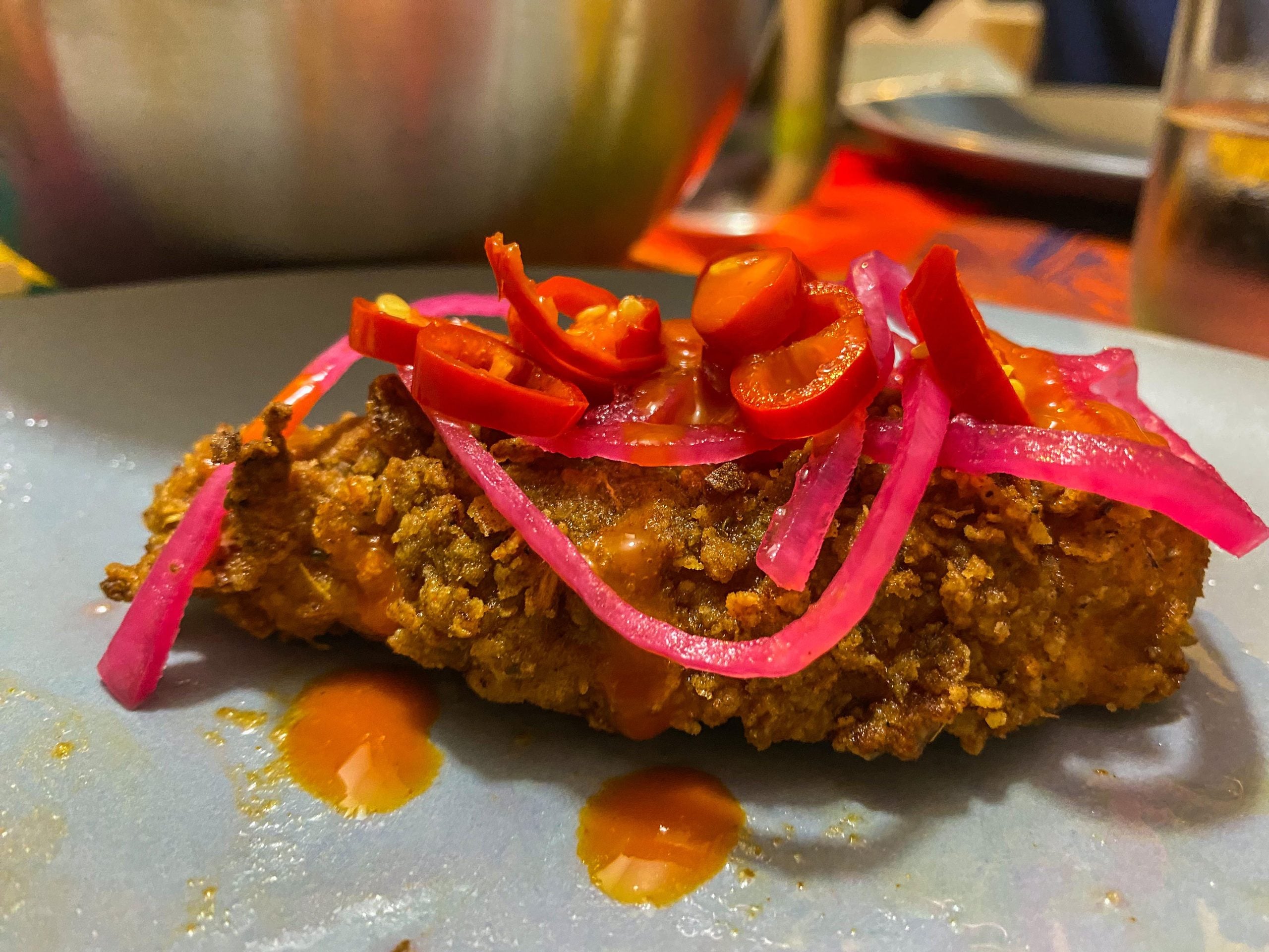 A piece of fried chicken drizzled with sauce and topped with sliced red chilies and pickled red onions.