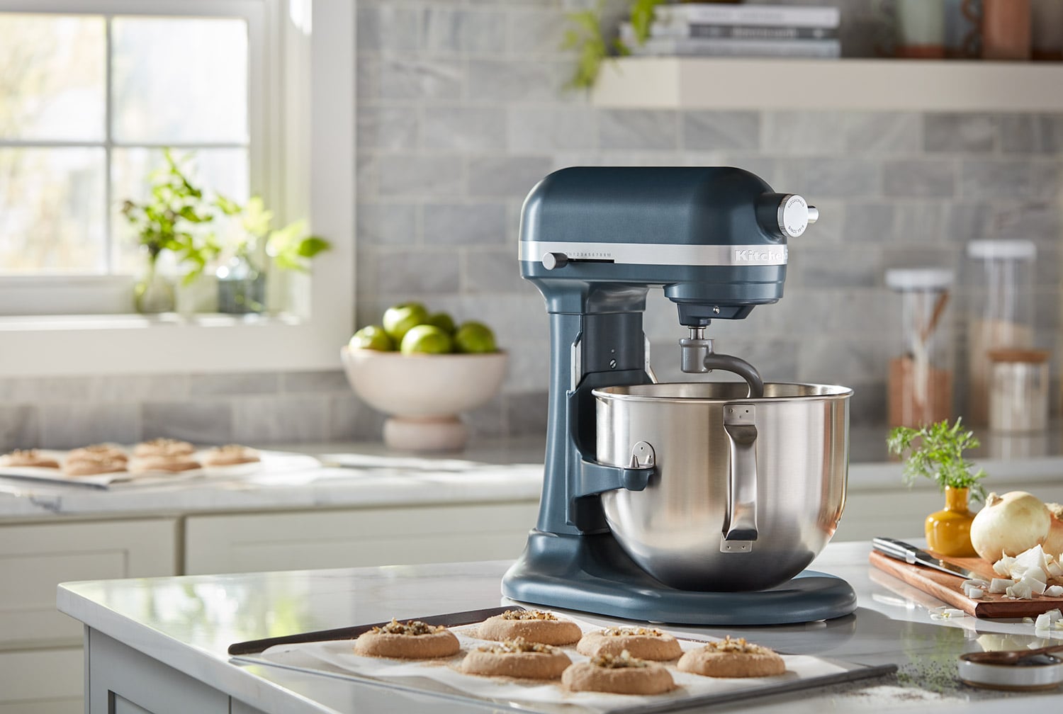 Dark blue KitchenAid stand mixer with a stainless steel bowl on a kitchen counter next to a tray of baked cookies.