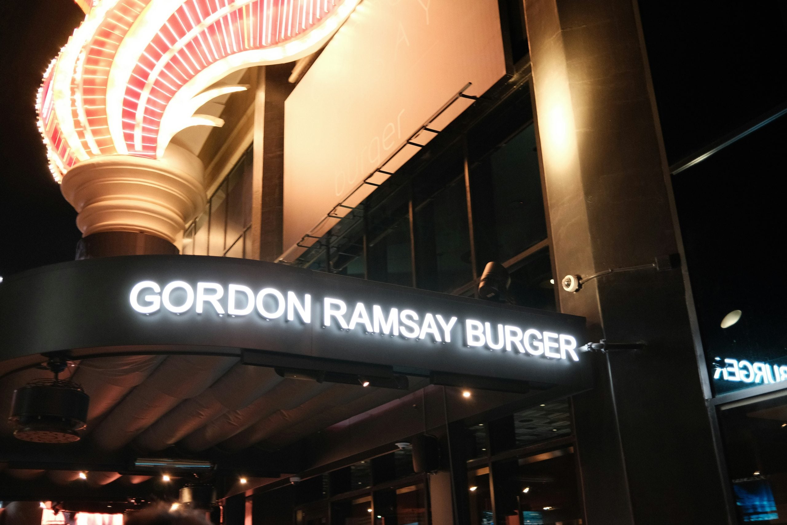 A brightly lit sign for “Gordon Ramsay Burger” is displayed on a building exterior at night, with decorative lights and reflections visible.