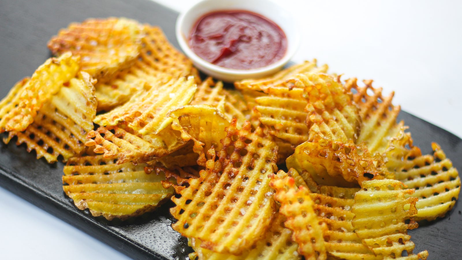 A pile of waffle-cut fries on a black serving board with a small bowl of ketchup.