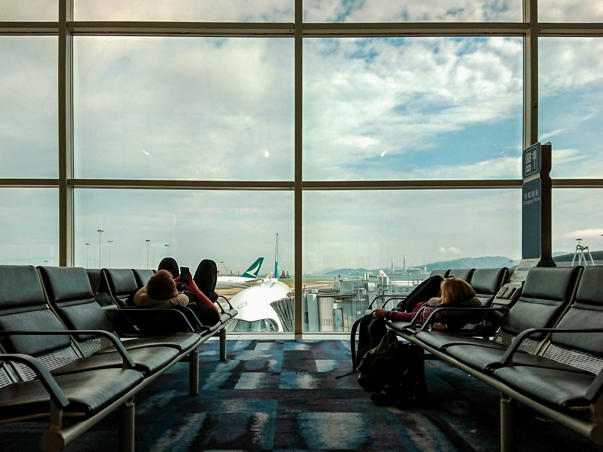 Two people lie across rows of black airport seats in front of a large window overlooking an airplane on the tarmac.
