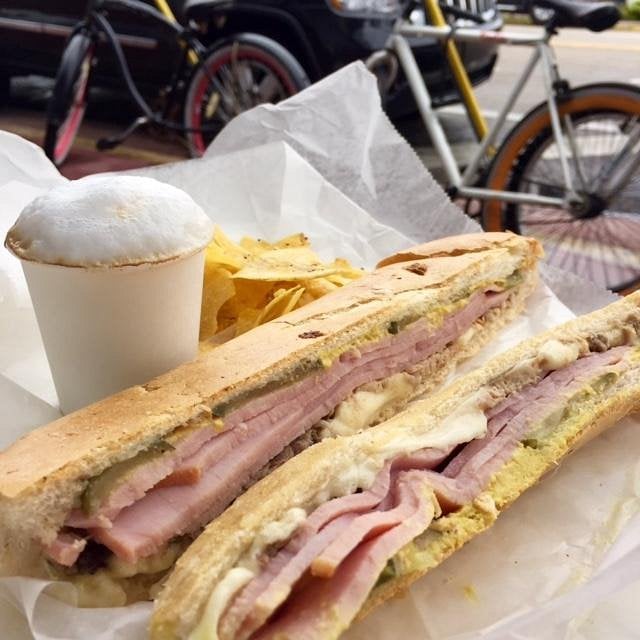 A pressed ham and cheese sandwich cut in half, served with a side of tortilla chips and a small cup of frothed coffee, with parked bicycles visible in the background.