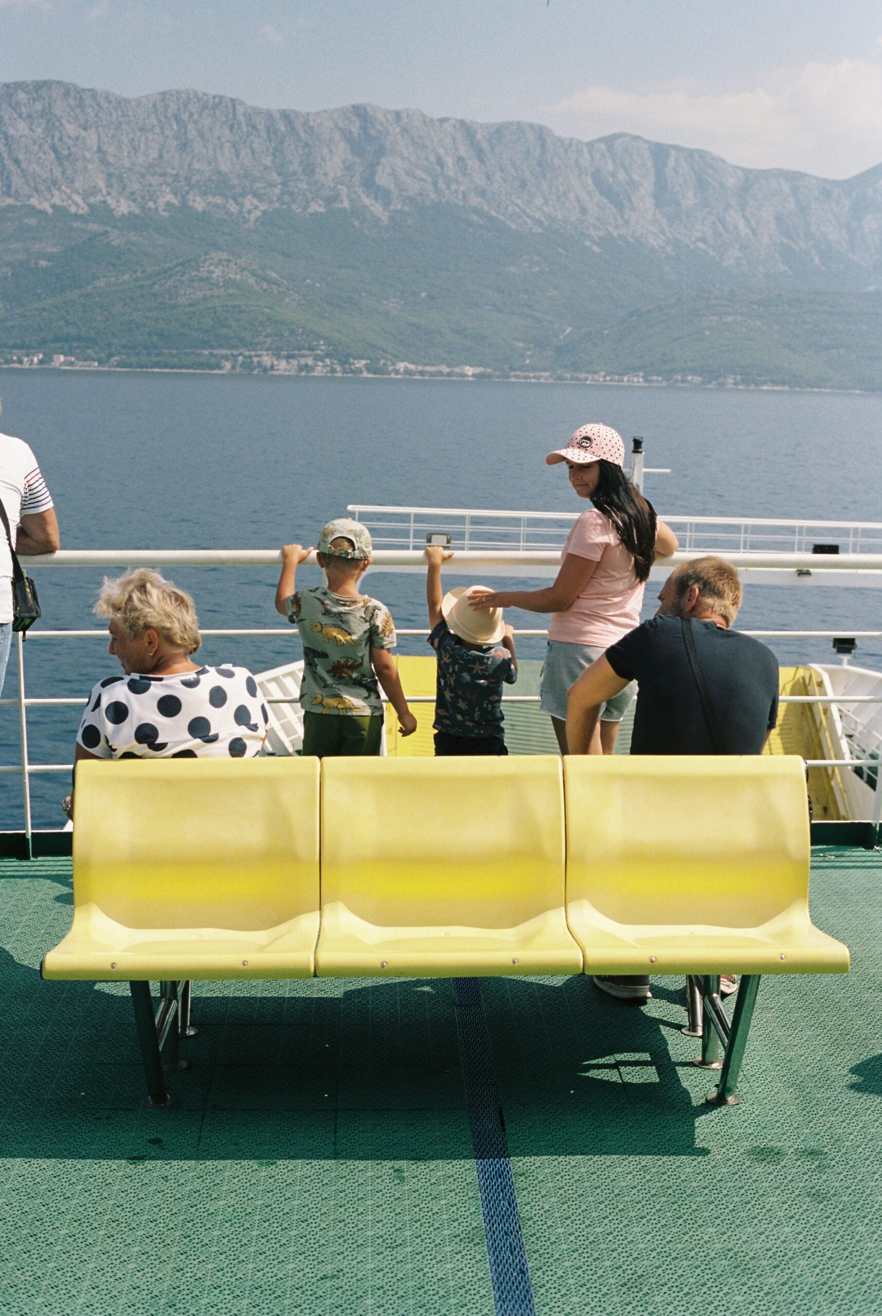 Five people sit and stand on a ferry deck with yellow seats, facing the sea and distant mountains. Two children stand at the railing, while adults sit or stand nearby, enjoying the view.