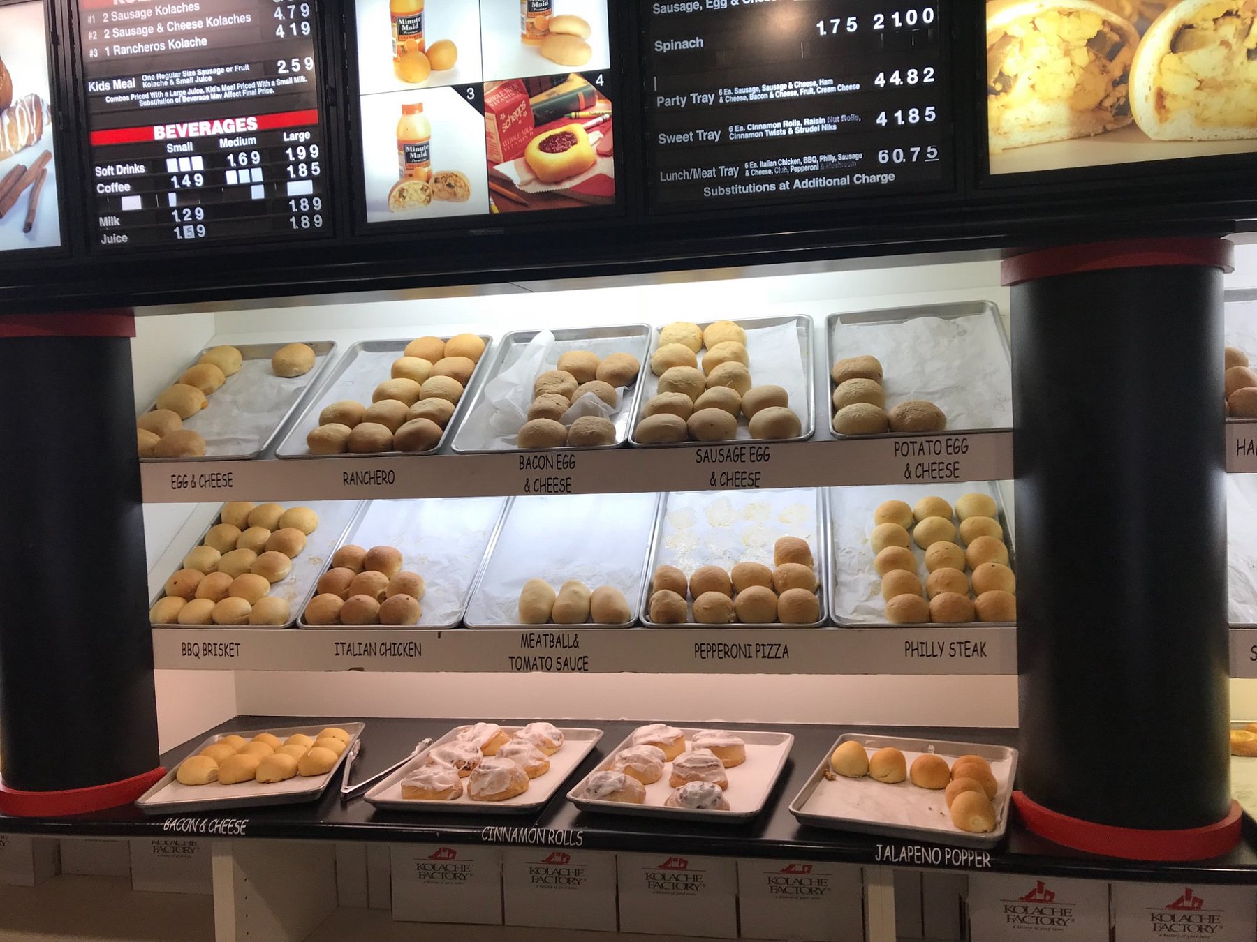 Display case at a Kolache Factory bakery featuring trays of various savory kolaches labeled by flavor, with menu boards above showing prices.