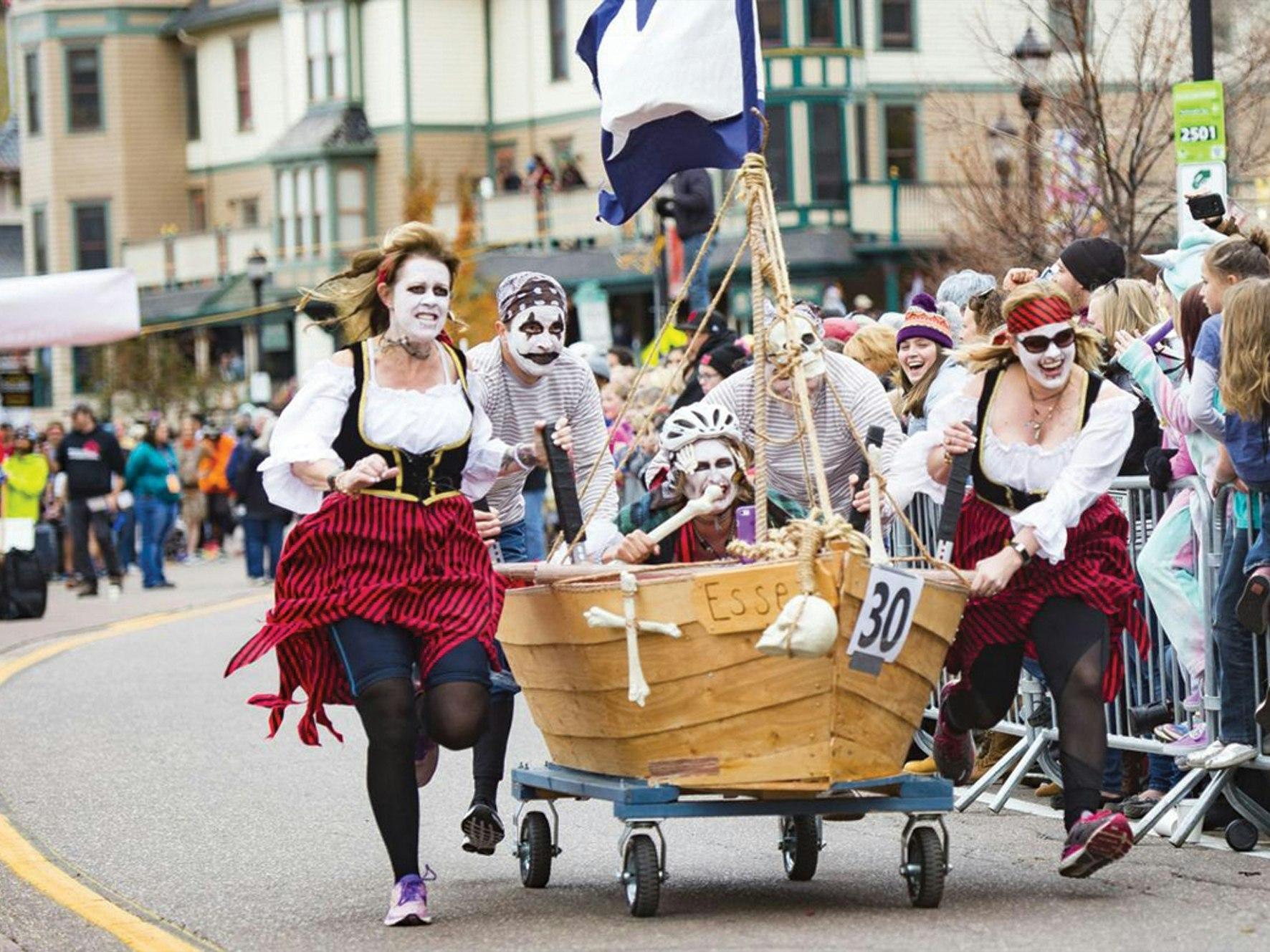 Four people dressed as pirates in face paint run down a street pushing a small, wheeled wooden boat decorated with a flag and the number 30.