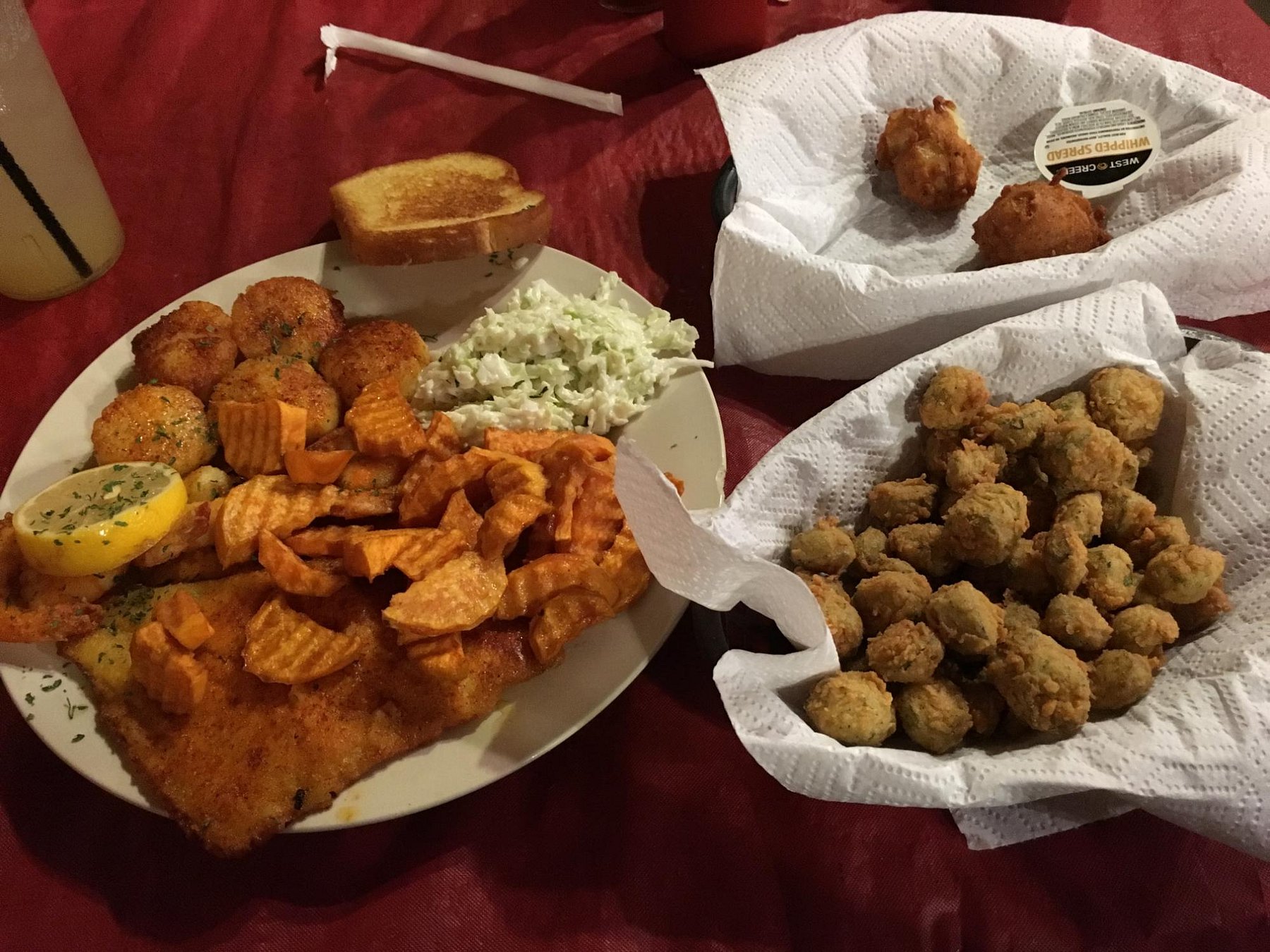 A plate of fried fish, sweet potato fries, hushpuppies, coleslaw, and toast, accompanied by two baskets of fried appetizers on a red tablecloth.