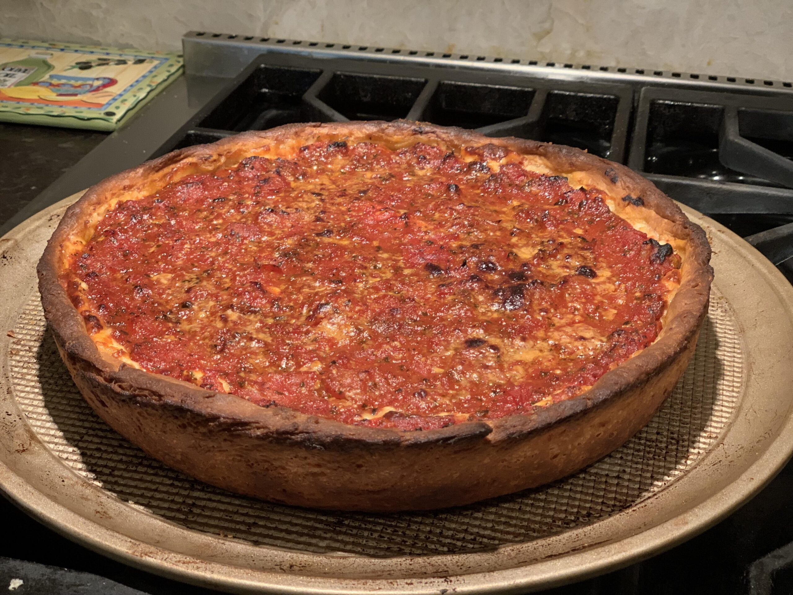 A baked deep-dish pizza with a thick, golden-brown crust and a layer of tomato sauce on top, sits on a round baking tray on a stovetop.
