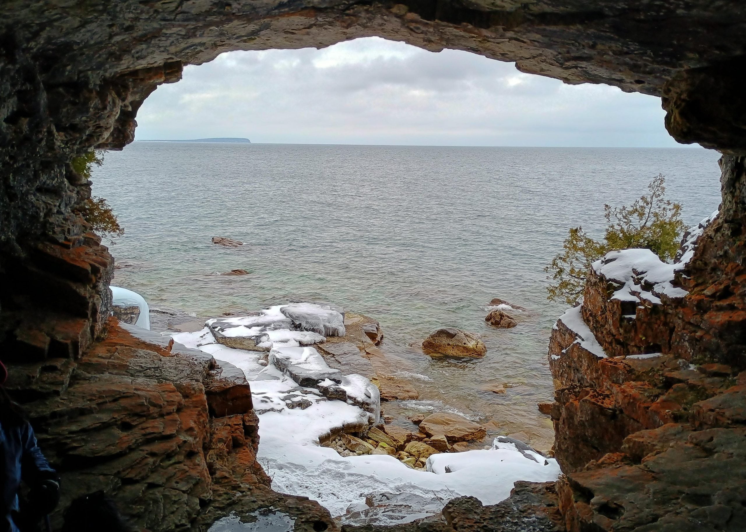 View of a lake and rocky shoreline seen from inside a dark stone cave, with patches of snow on the ground.