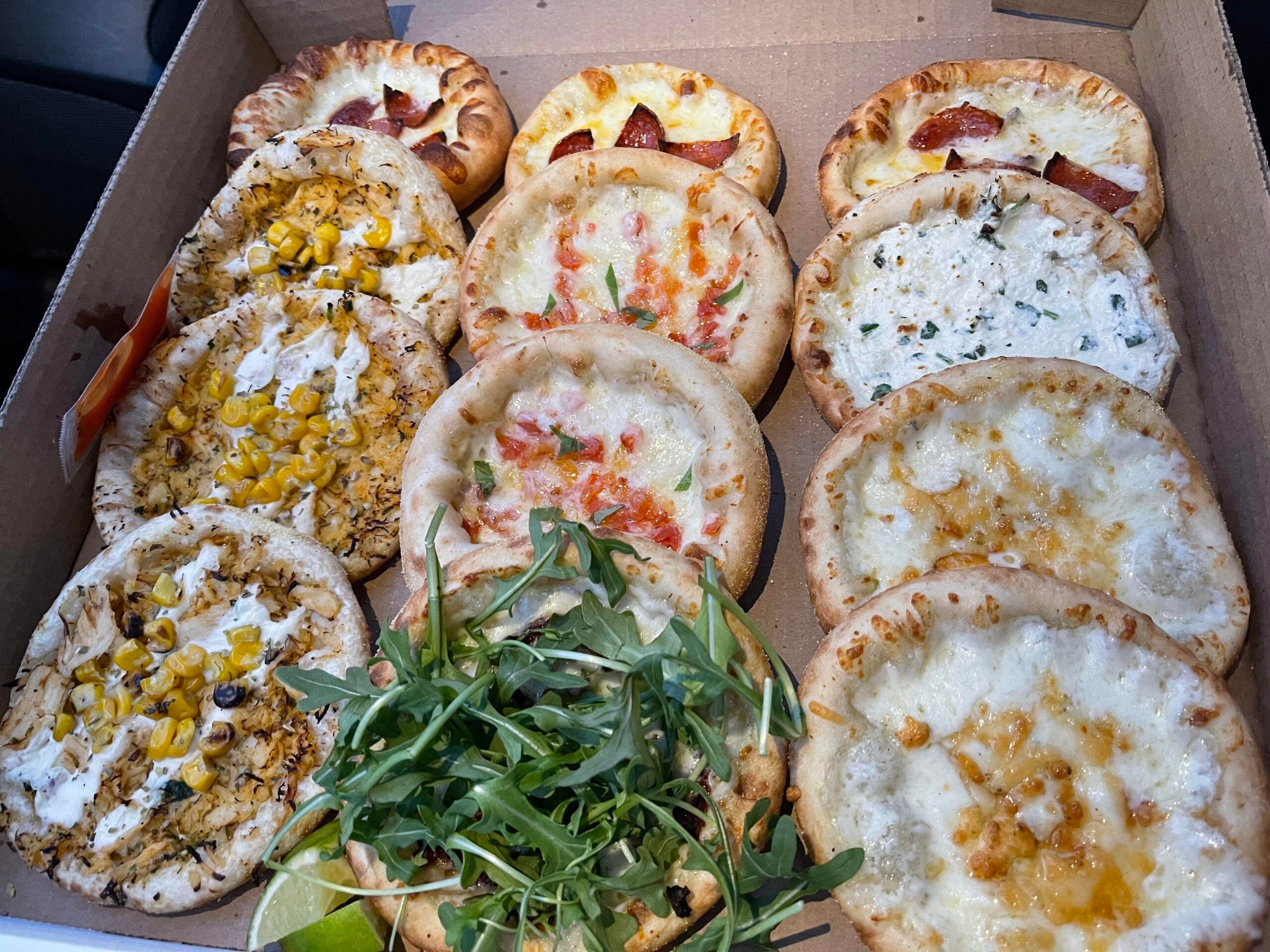 A cardboard box filled with ten assorted mini pizzas topped with ingredients including corn, cheese, pepperoni, herbs, and fresh arugula.