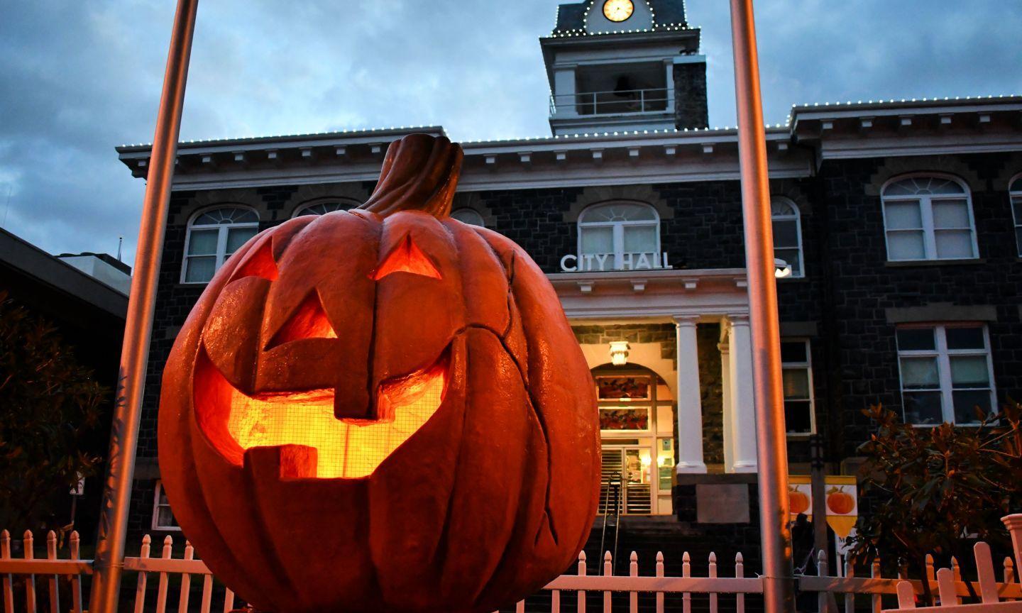A large, illuminated jack-o’-lantern sits in front of a stone City Hall building at dusk.
