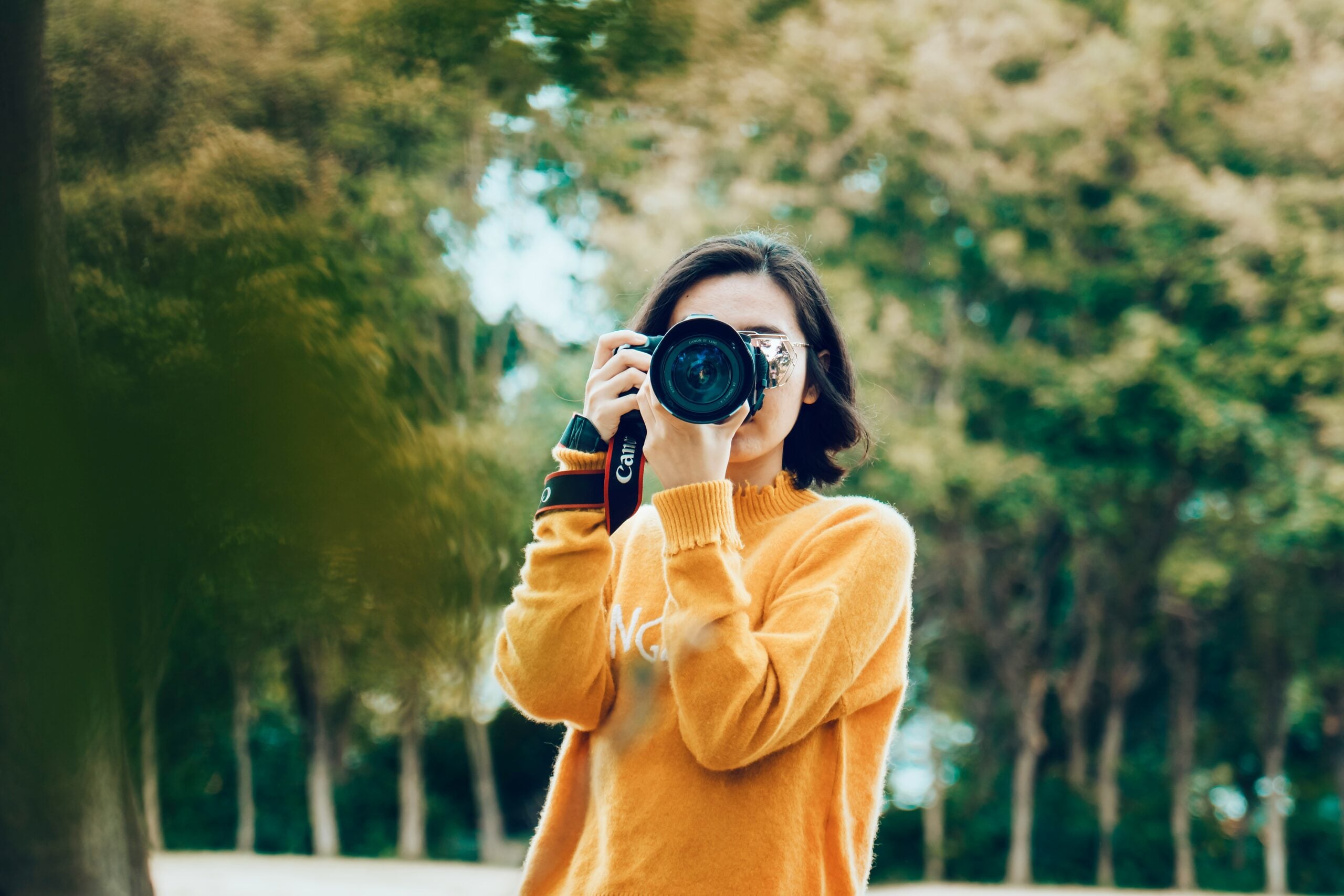 A woman in a mustard yellow sweater holds a camera to her eye to take a picture outdoors.