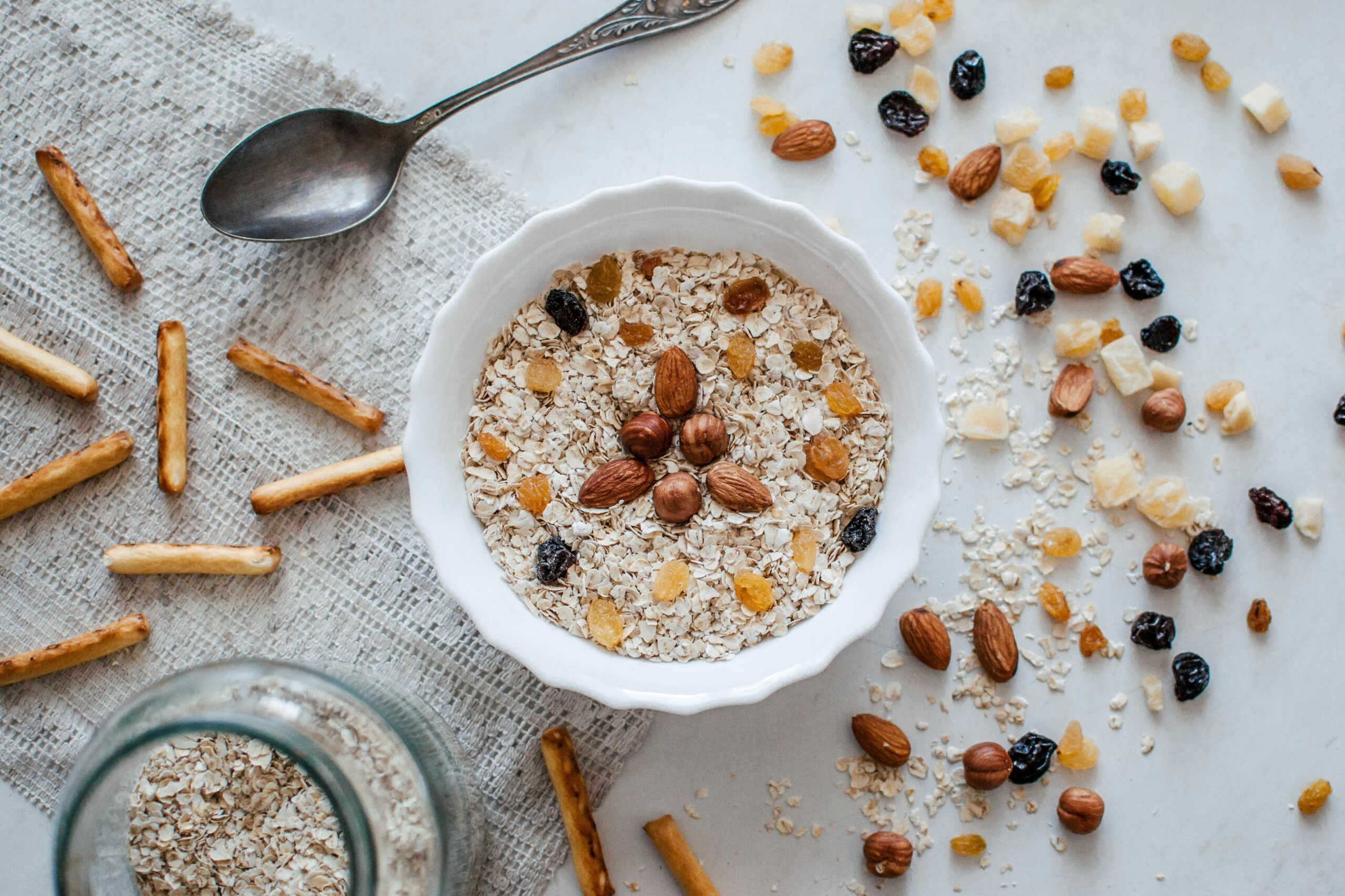 A white bowl of oatmeal topped with almonds, hazelnuts, raisins, and dried fruit sits on a cloth next to a spoon, scattered granola, pretzel sticks, and a glass jar of oats.