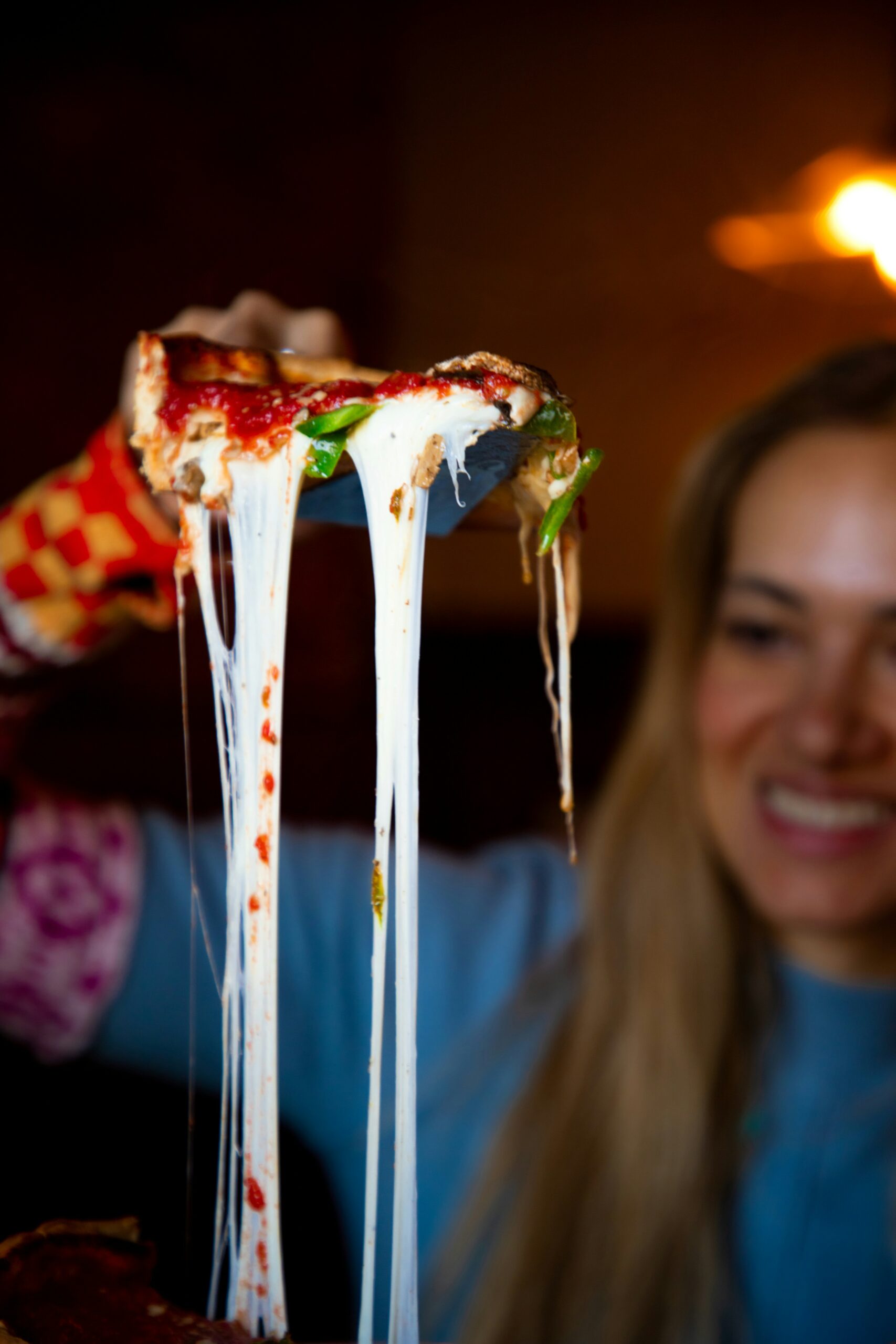 A person lifts a cheesy slice of pizza, with melted cheese stretching, while smiling in the background.