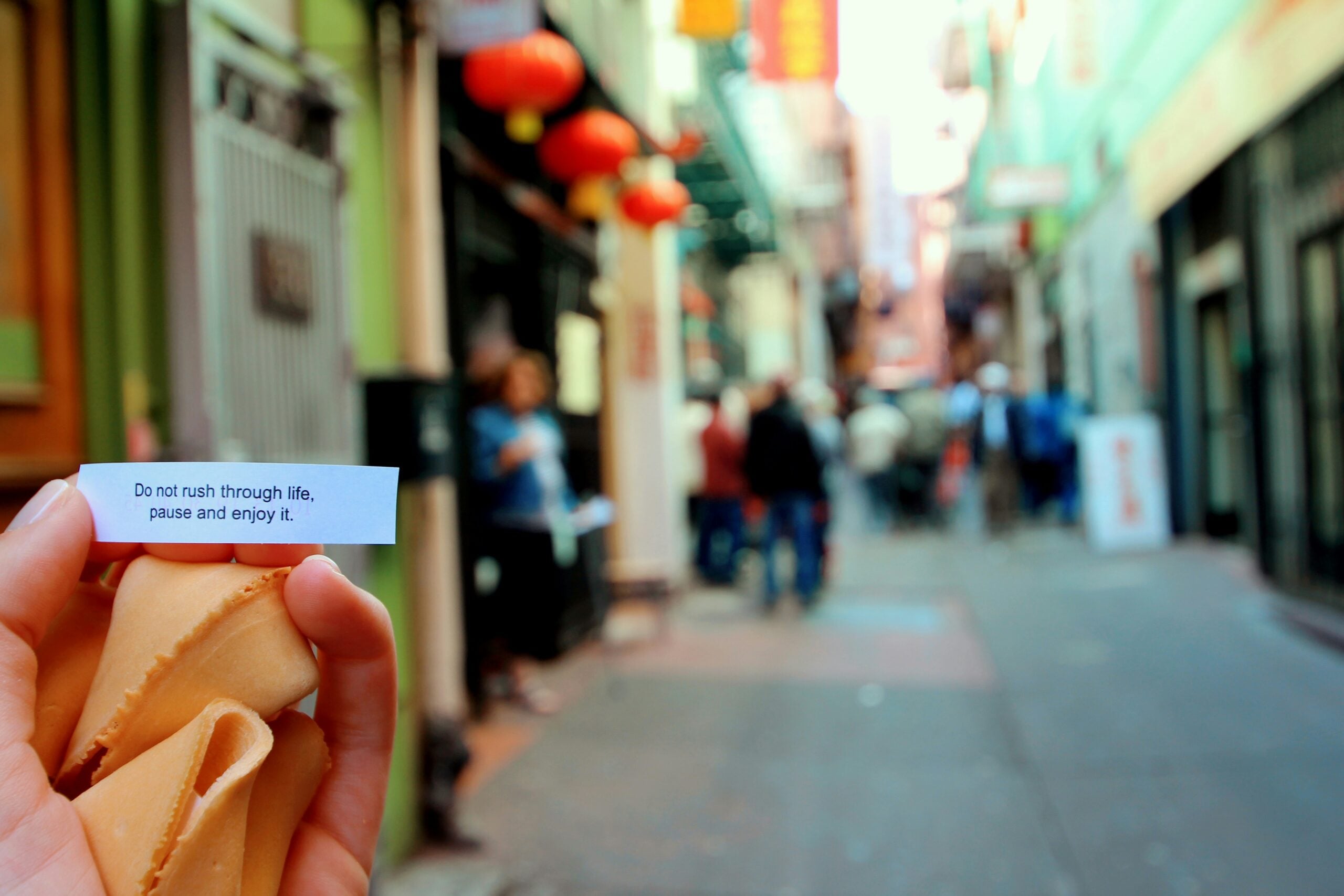 A hand holding a fortune cookie and a small slip of paper that reads, “Do not rush through life, pause and enjoy it,” in front of a blurred street scene.