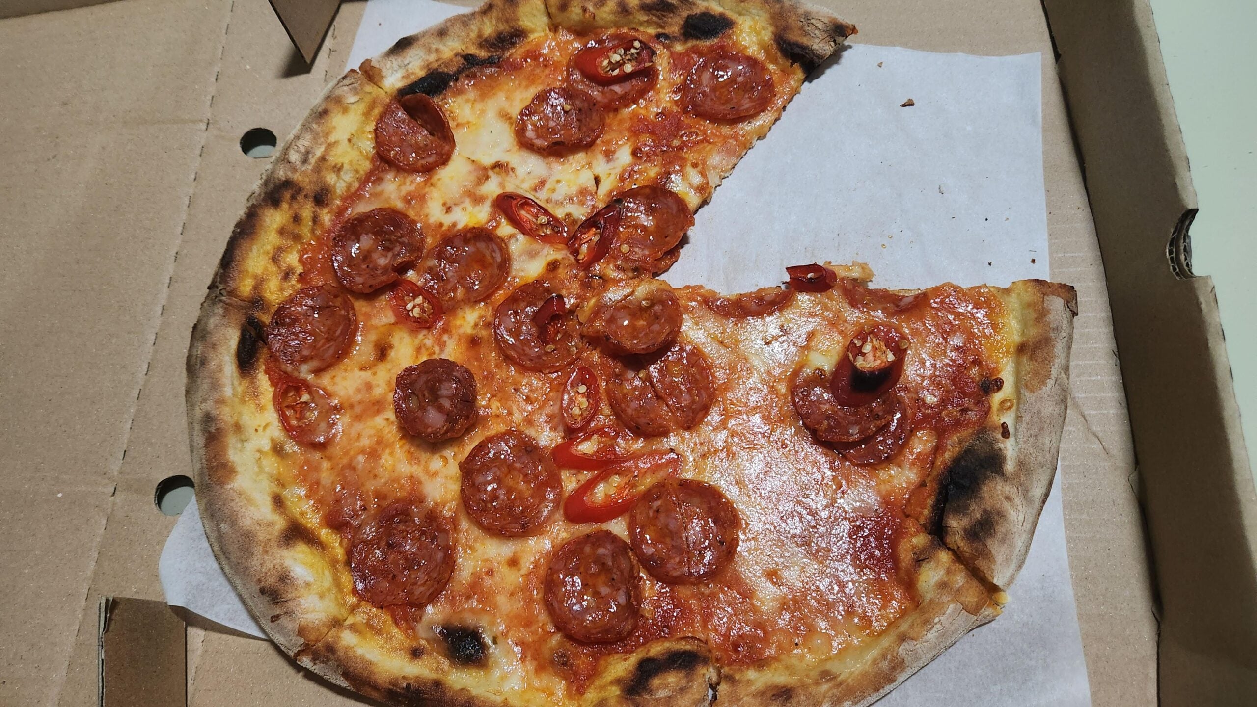 A pepperoni and chili pizza in a cardboard delivery box, with one slice missing.
