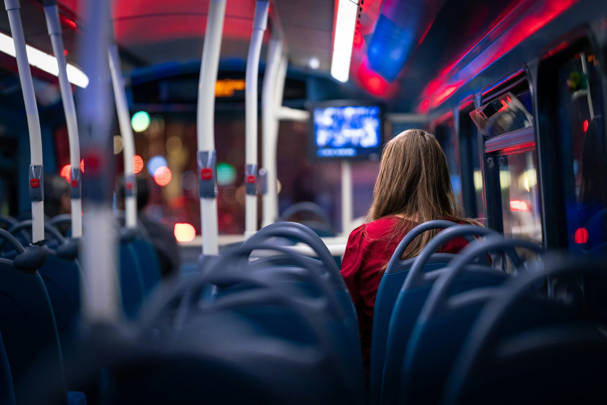 A person with long hair wearing a red top sits in a blue bus seat at night, facing the front of the vehicle.