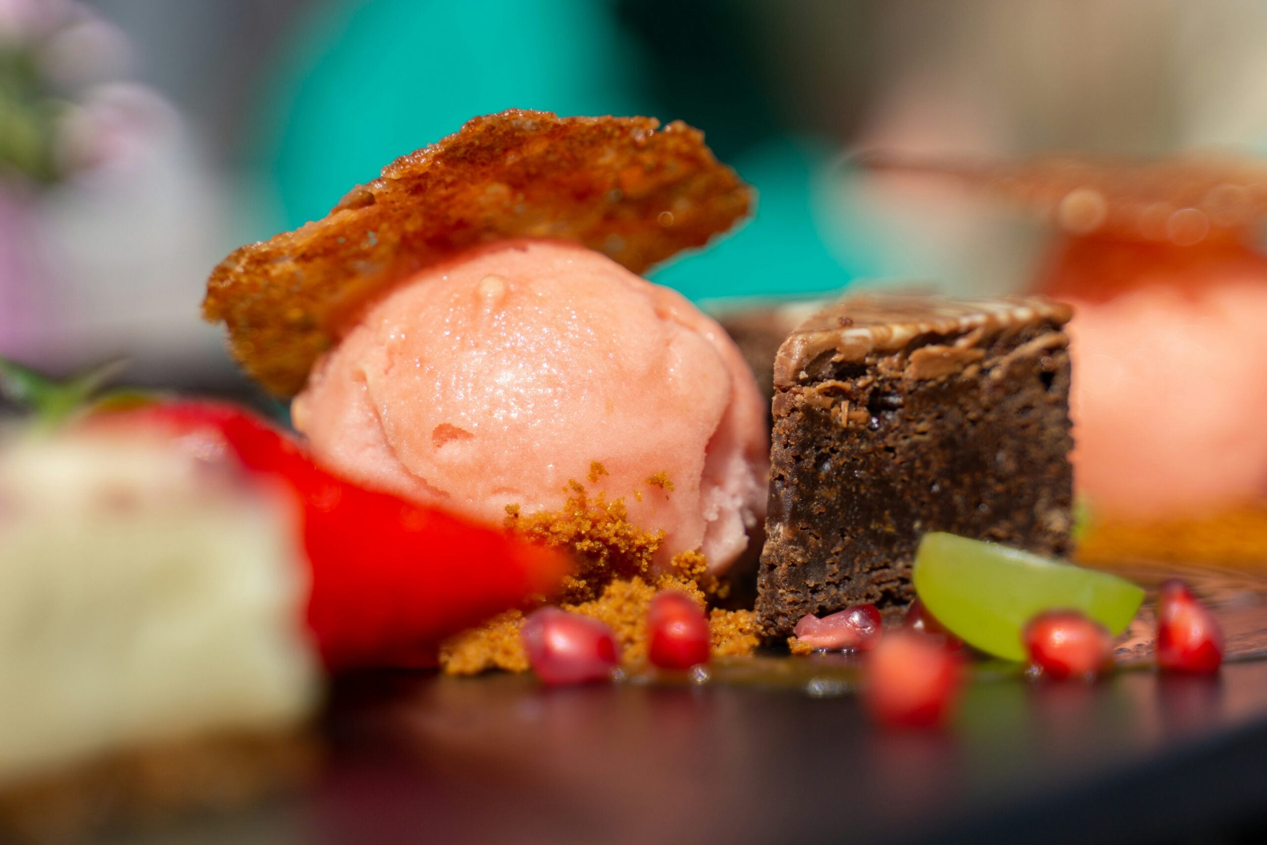 A scoop of pink sorbet topped with a crisp wafer, served next to a chocolate brownie, pomegranate seeds, and a green grape.