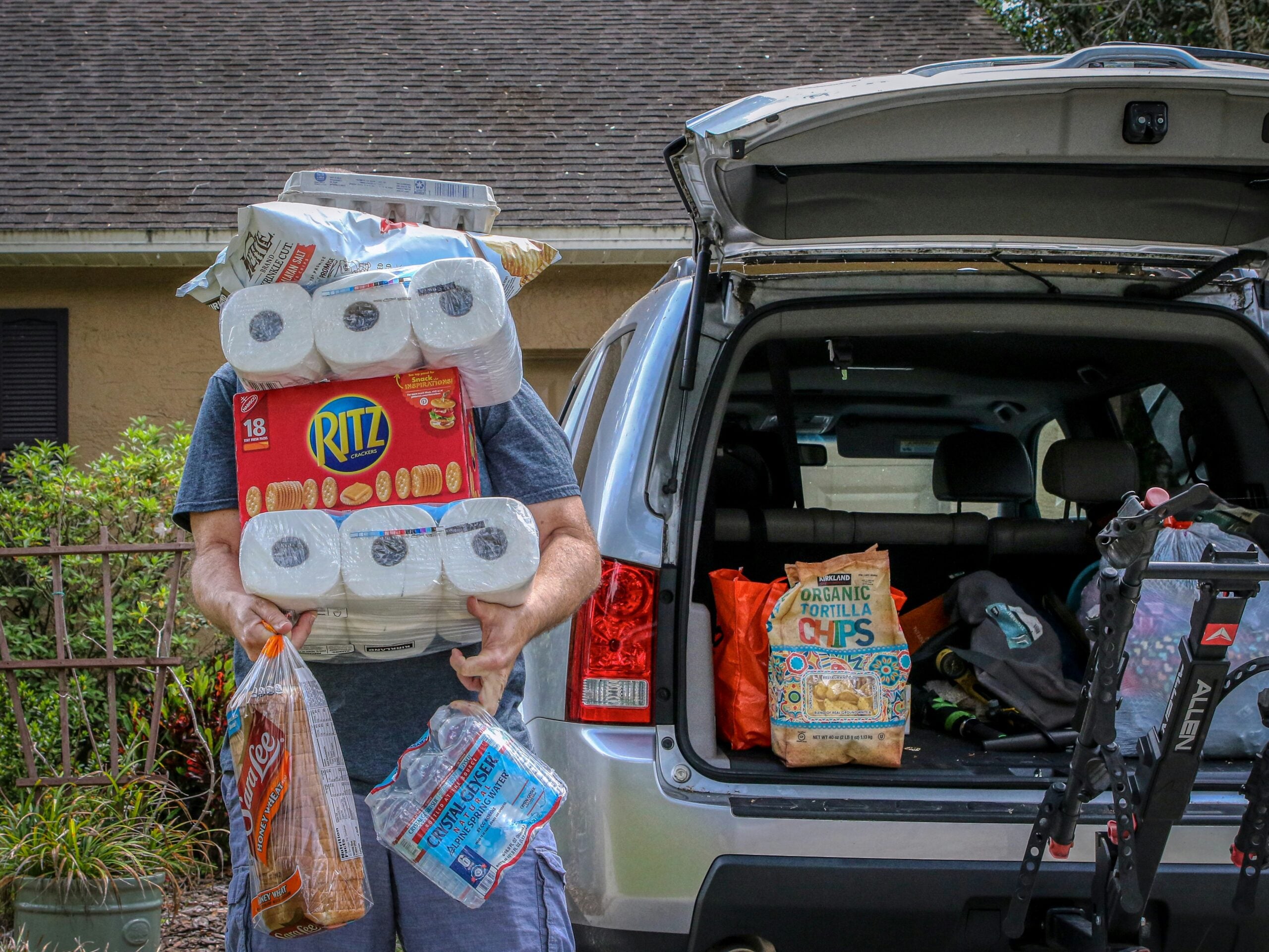 A person carries a large stack of groceries, including toilet paper, crackers, bread, and water, in front of an open car trunk containing more bags.