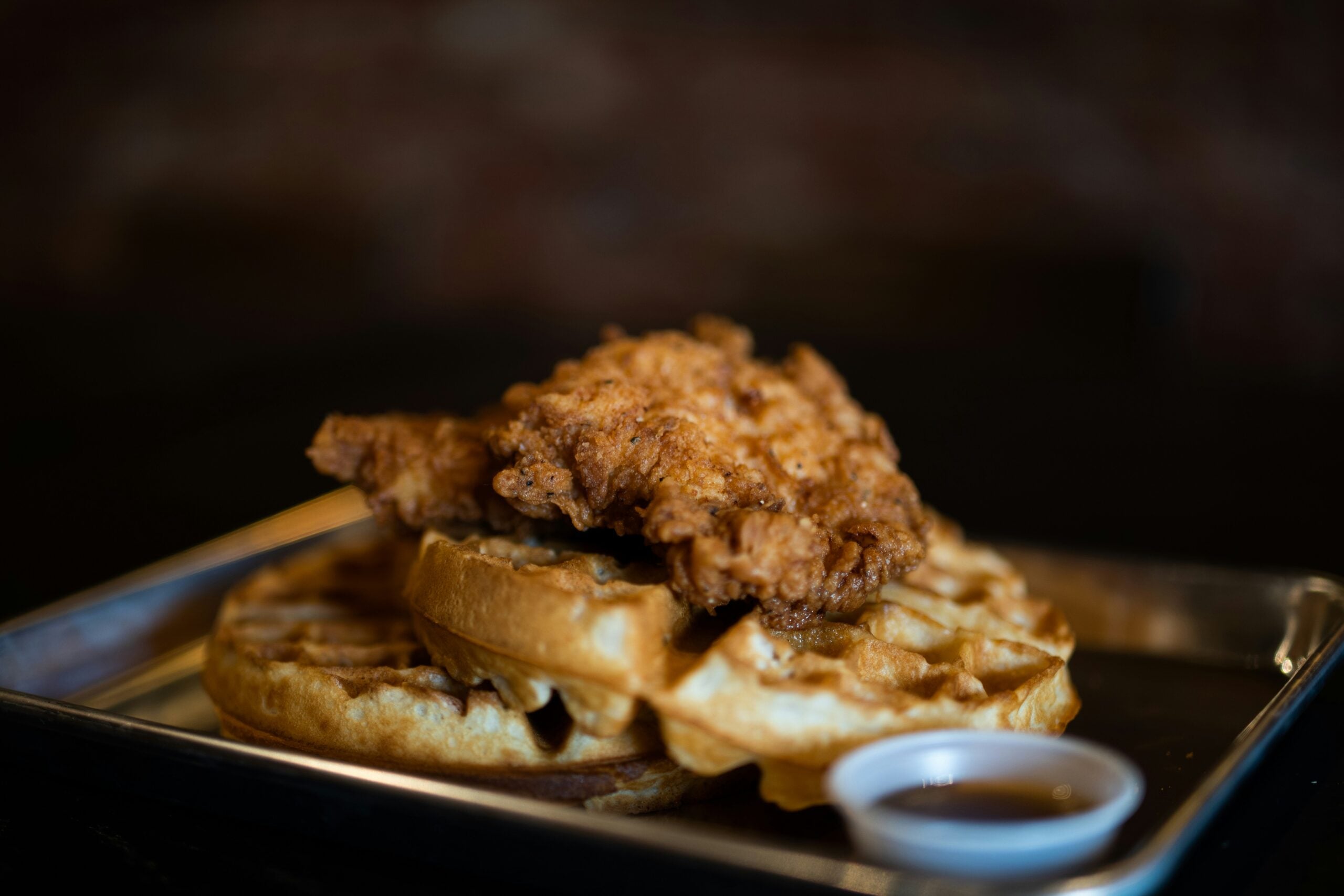 Fried chicken on top of a Belgian waffle served on a metal tray with a small container of syrup.