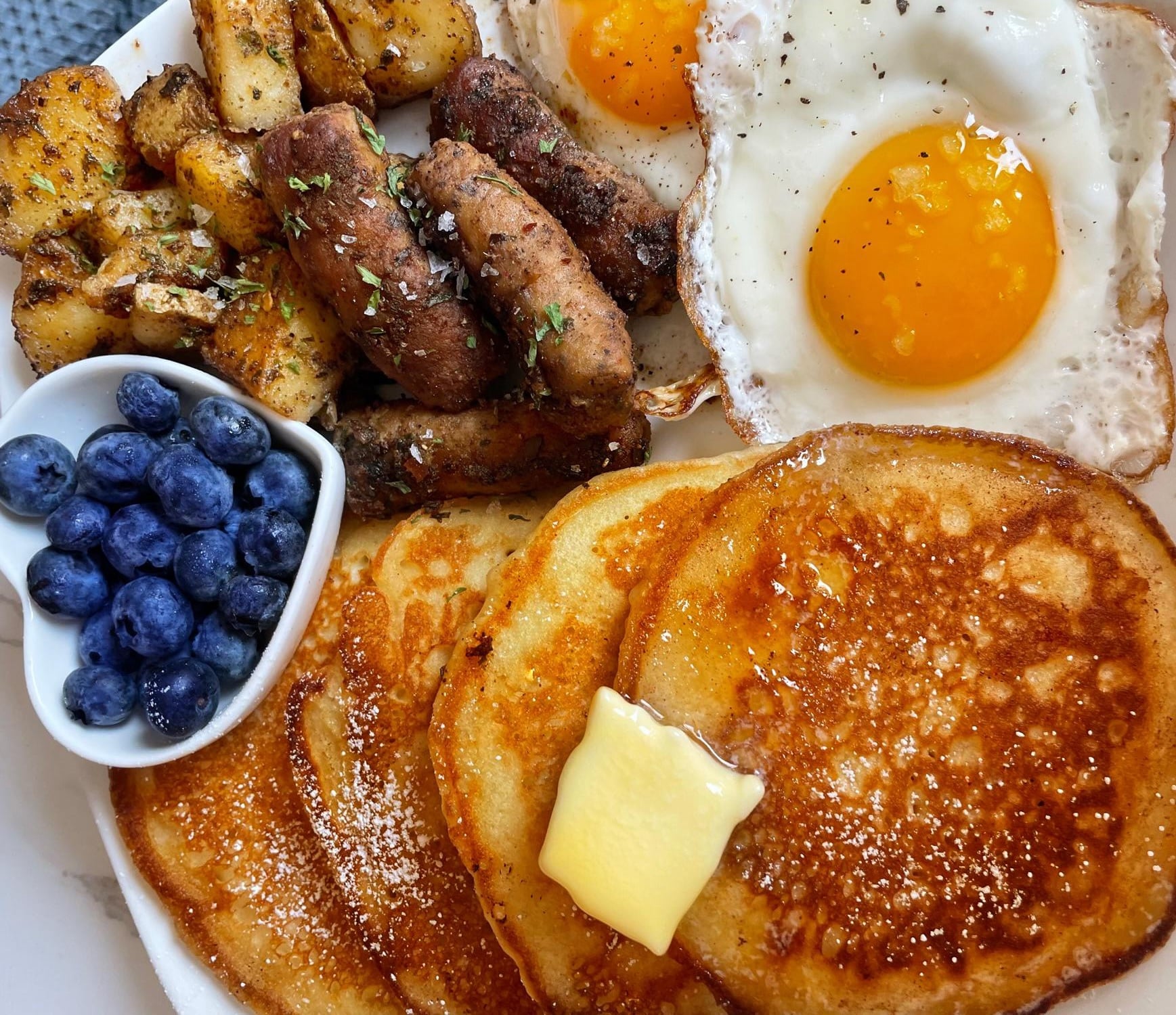 Breakfast plate with pancakes topped with butter, two fried eggs, breakfast sausages, seasoned potatoes, and a small heart-shaped bowl of blueberries.